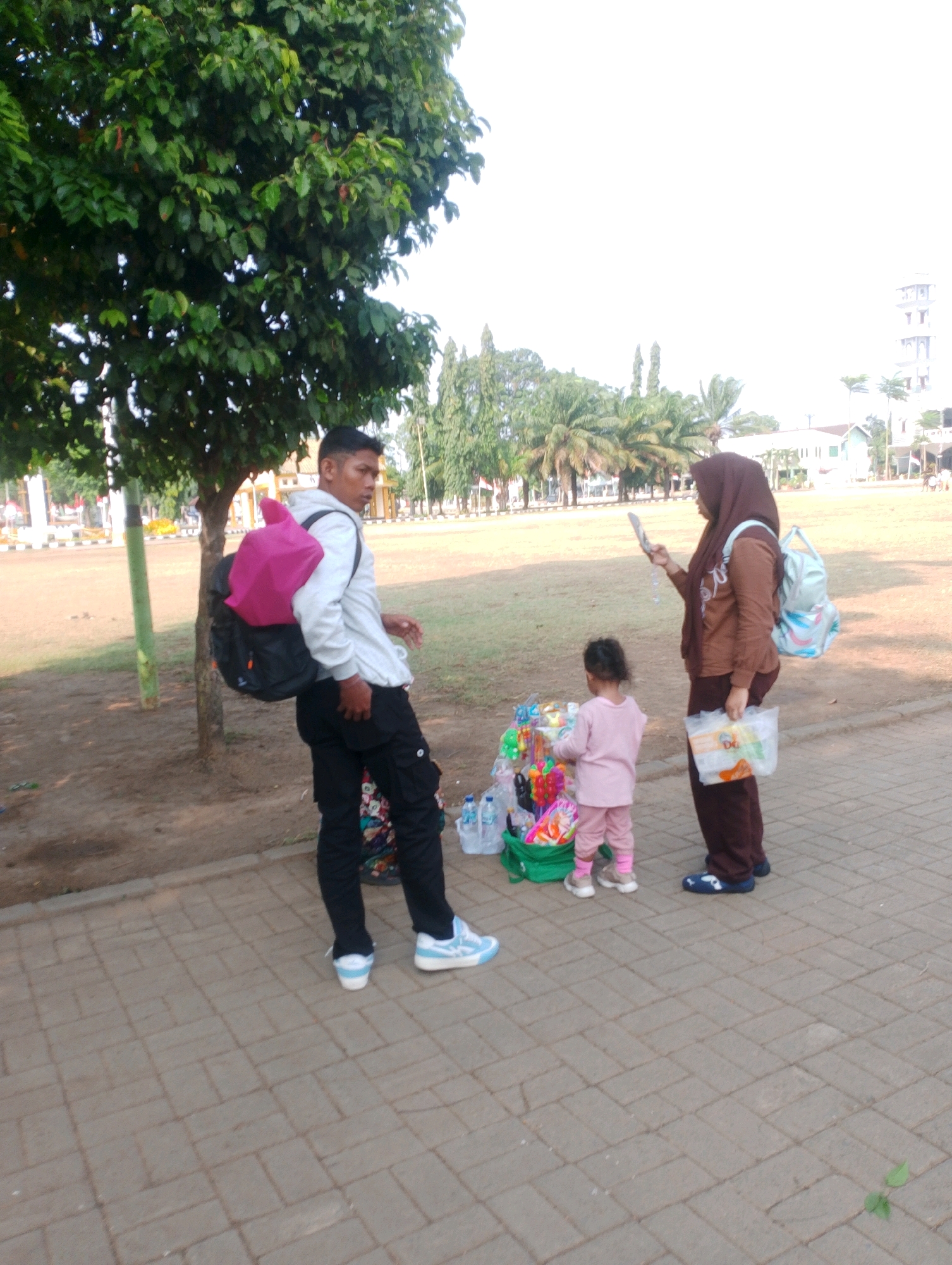 A young man wearing a white hoodie, black cargo pants, and blue and white sneakers stands on a brick-paved pathway. He has a large black backpack on his back, and a bright pink backpack is resting on top of it. To his right, a woman in a brown hijab and brown clothing is holding a mirror and looking at it. She has a light blue backpack with a pattern on it. In front of the woman, a toddler in a pink outfit is looking at a small cart filled with toys and plastic bags. The cart is green. Behind the people, there is a grassy area with some trees and palm trees. In the background, there are buildings and a tall tower. The scene appears to be outdoors, possibly in a park or public square in Probolinggo, Indonesia, given the presence of palm trees and architectural styles. The lighting suggests it is daytime.