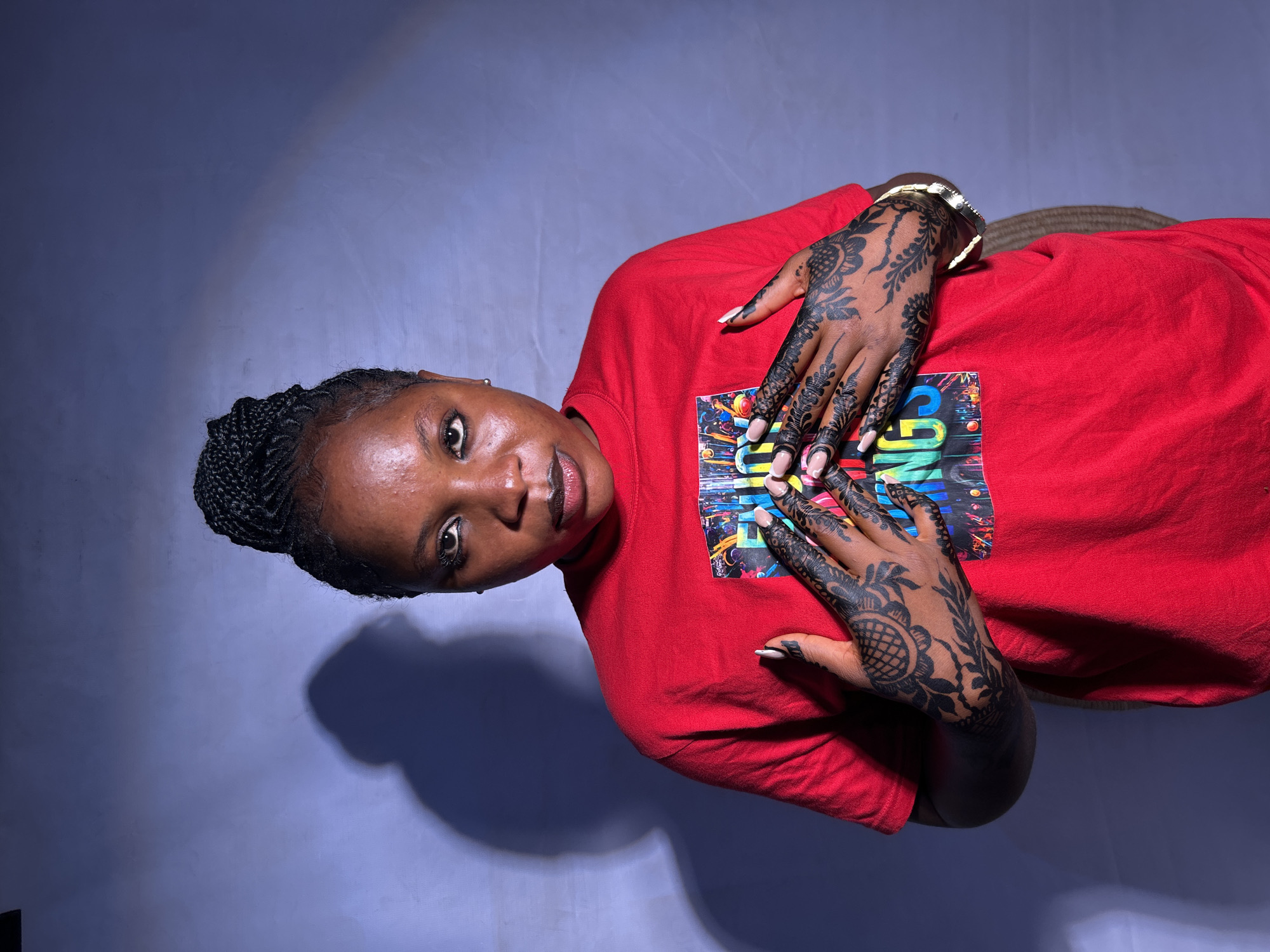 A portrait shot from slightly above shows a woman with dark hair braided on top of her head. She is wearing a bright red t-shirt. Both of her hands are visible, adorned with elaborate henna designs. Her nails appear to be manicured with white tips. The background is a gradient of grey and blue. The location within Jos, Nigeria is not determinable from the image.