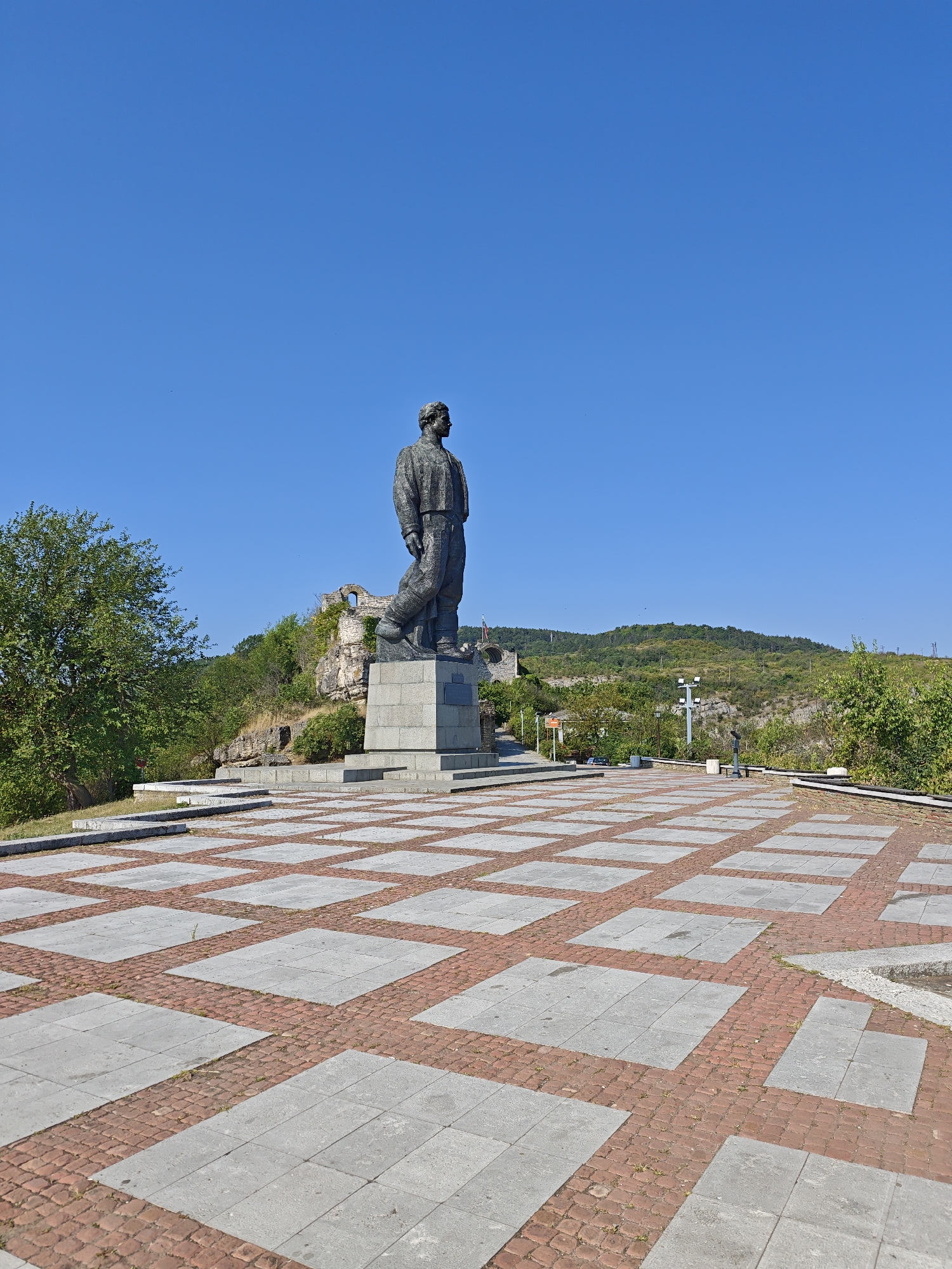 The image depicts the monument of General Vasili Levski in the city of Lovech, Bulgaria. The monument is a large bronze statue of a man standing on a stone pedestal, which is set within a wide, paved plaza. The plaza is laid with a geometric pattern of large, gray square tiles interspersed with smaller, reddish-brown bricks. In the background, behind the monument, are the ruins of an ancient fortress, a rocky cliff face covered in vegetation, and a green, hilly landscape under a clear blue sky. There are no people visible in the image. The overall scene is one of a historical landmark situated in a natural, hilly environment.