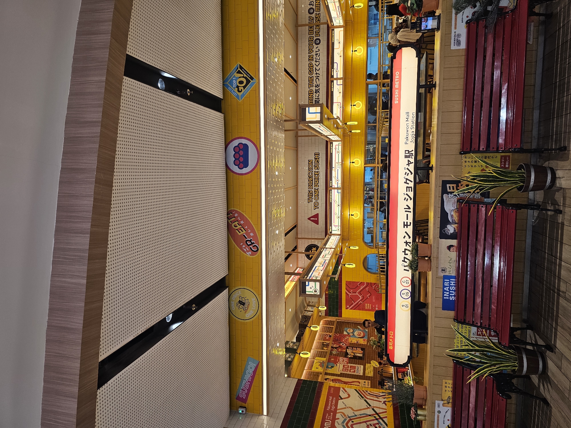 The image shows the exterior of a brightly lit sushi restaurant with a yellow tiled facade. A long, illuminated counter with "SUSHI METRO" signage is prominently displayed. The signage also features Japanese characters and "Pakuwon Mall Jogja Station" in English. Several decorative elements, including circular signs with Japanese text and graphics, are visible along the upper yellow wall. Red benches are placed in front of the counter, with potted plants on either side. In the background, through the windows of the restaurant, customers and staff can be seen. The overall impression is a modern, vibrant dining establishment, likely situated in a shopping mall or public area in Condongcatur, Indonesia, given the mention of Pakuwon Mall.