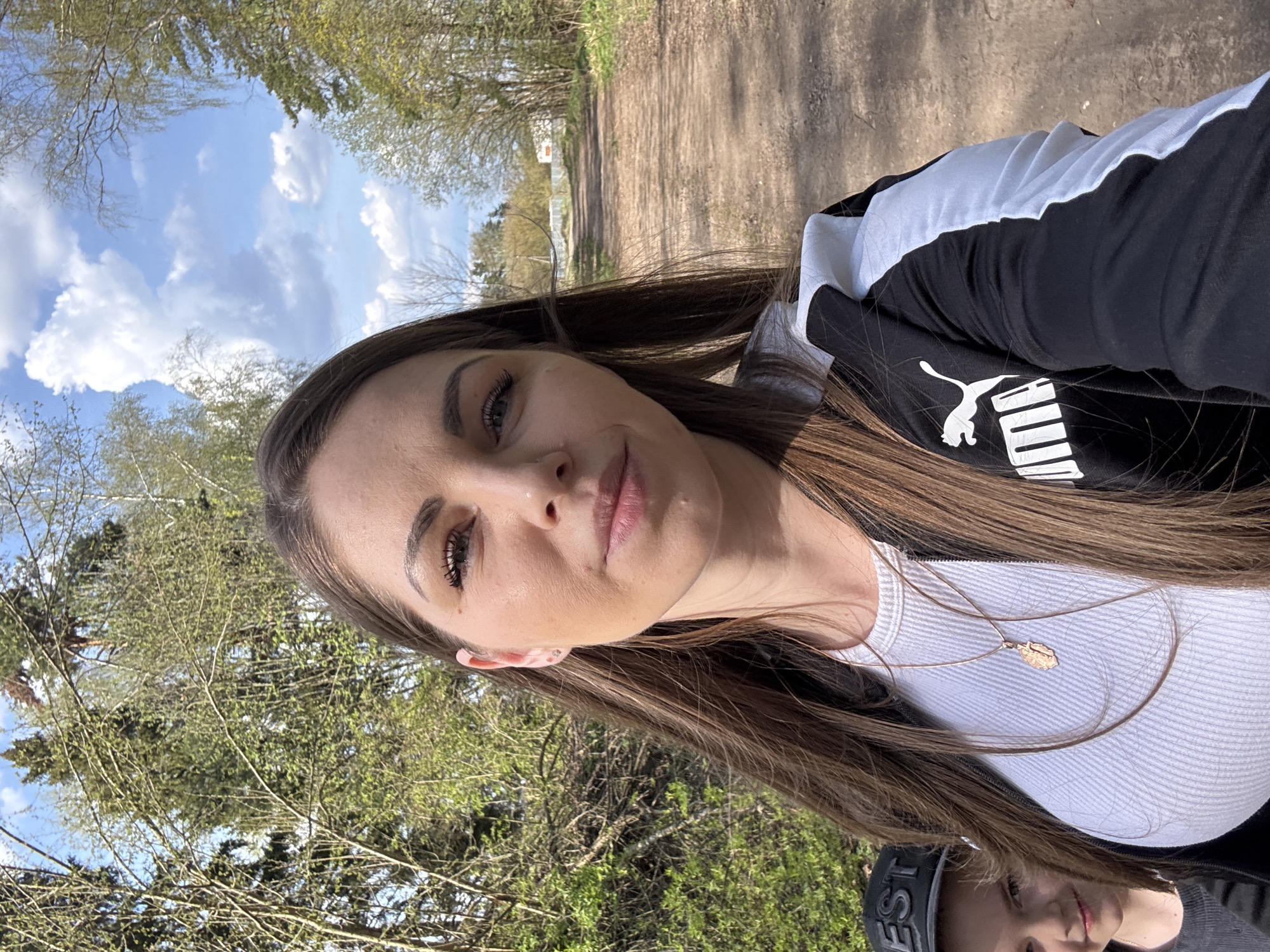 A woman takes a selfie outdoors in Раздолье, Russia. She has long brown hair, a white ribbed shirt, and a black Puma jacket. She wears a gold necklace. The background shows a dirt path lined with trees, a white building, and a partly cloudy sky. A second person is partially visible in the bottom right corner of the frame, wearing a grey shirt and a baseball cap.