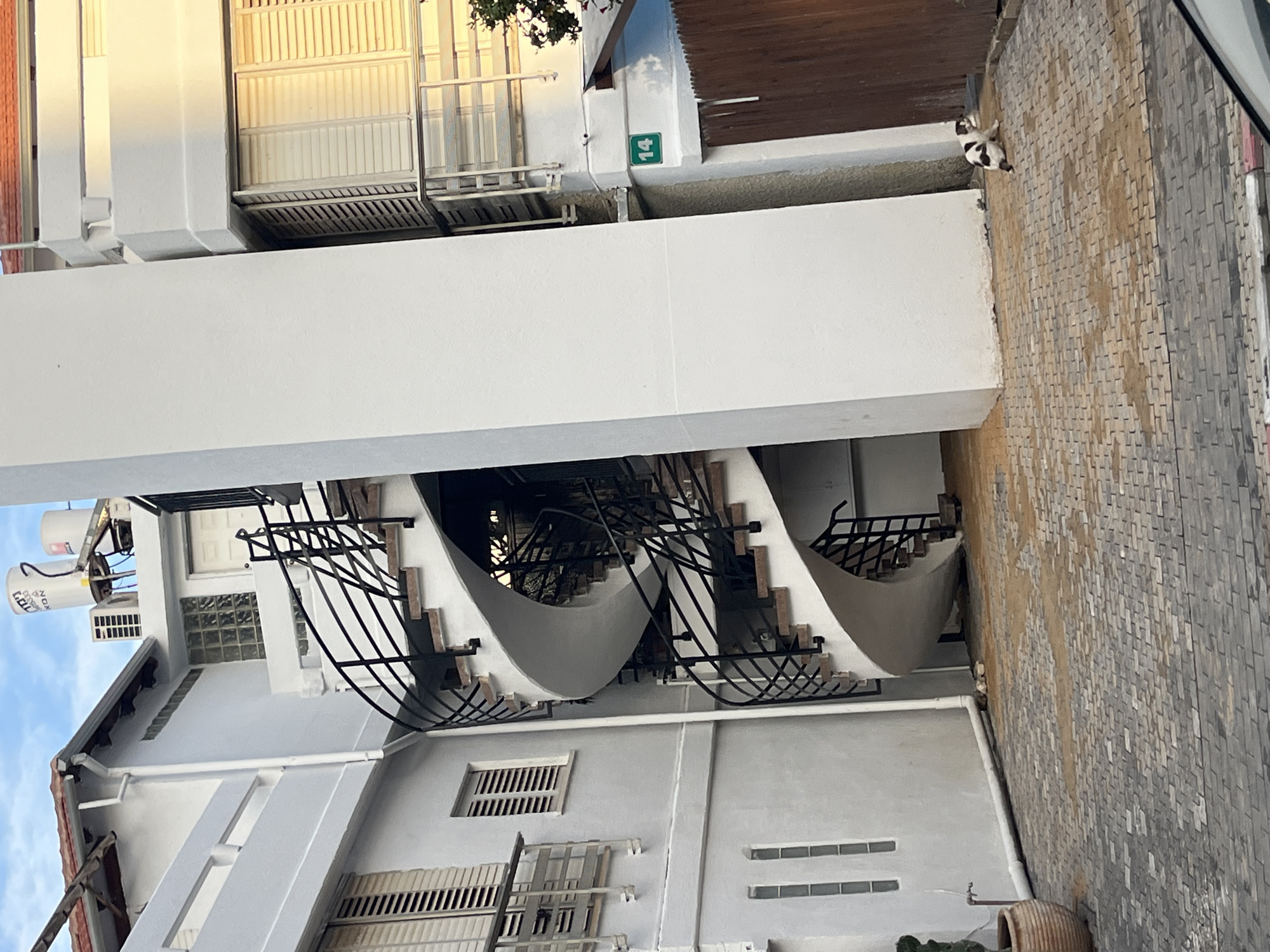The image displays the exterior of a multi-story white building located in Ramat Gan, Israel. A prominent white concrete spiral staircase with dark brown risers and black metal railings is centrally positioned, ascending against the building's facade. To the left, a portion of the building shows multiple windows with closed white louvered shutters, several balconies with light-colored railings, and a vertical white downspout. A terracotta pot is visible at ground level near the bottom left. The right side of the structure features a substantial white vertical column, a green square sign with the white numeral "14", and a section of a wooden fence. The rooftop includes terracotta tiles, white gutters, air conditioning units, and two white cylindrical water heaters/tanks, one bearing Hebrew text. The ground area is a paved courtyard composed of gray and brown interlocking pavers, showing widespread brown staining or discoloration. A small, short-haired cat, primarily white with black patches, is seated on the paving near the wooden fence, facing away from the viewer. The scene is illuminated by natural light under a blue sky with scattered clouds. No human subjects are present.