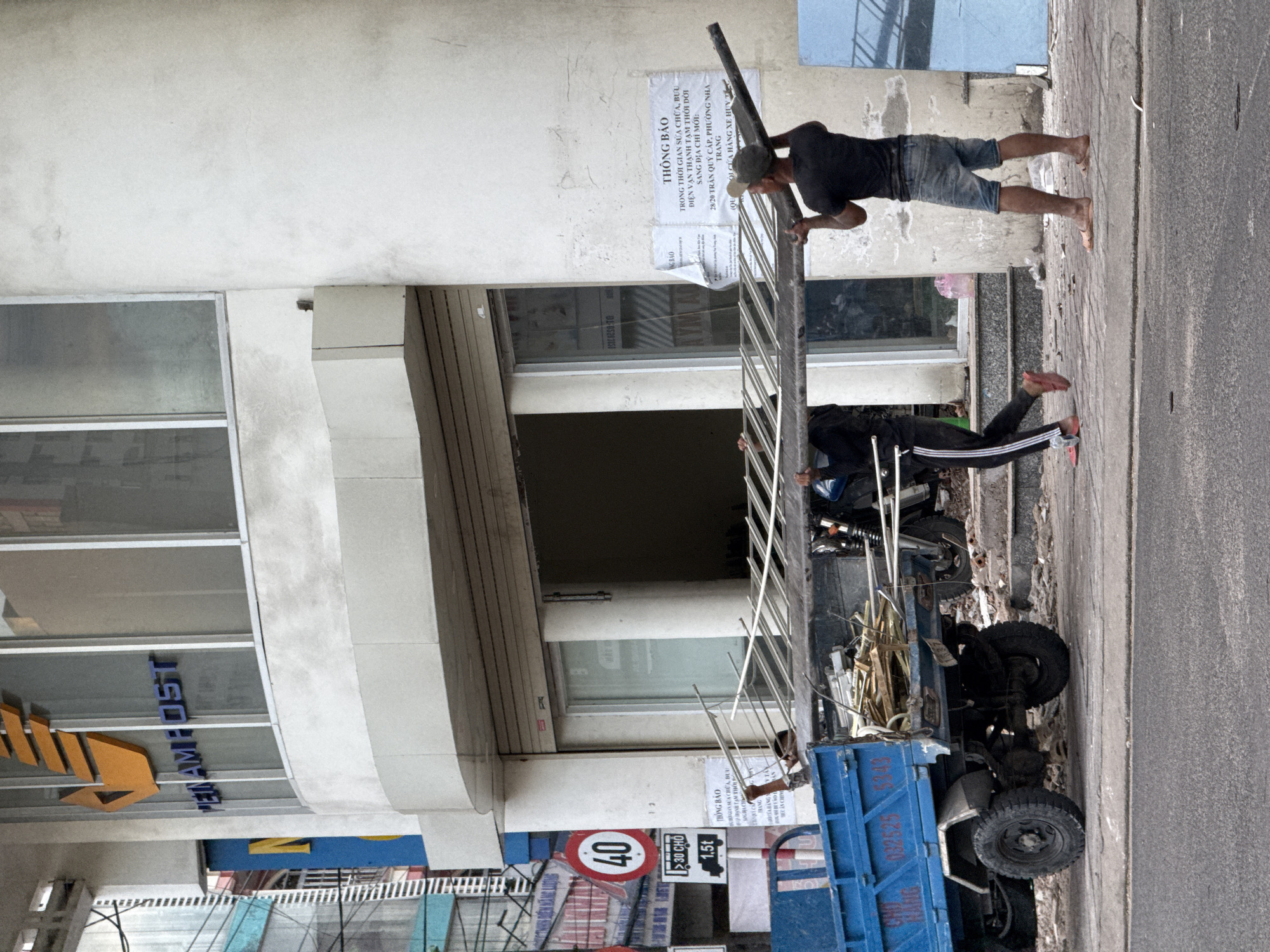 Two men are carrying a long metal frame in front of a building in Nha Trang, Vietnam. The building has a large sign that reads "VIETNAMPOST." The metal frame is being carried horizontally, with one man holding the front and the other the back. They are walking on a paved sidewalk. To the left of the building, a blue truck is parked. The truck is loaded with building materials. There are traffic signs on the side of the road, including a speed limit sign of 40. The scene appears to be in an urban environment.