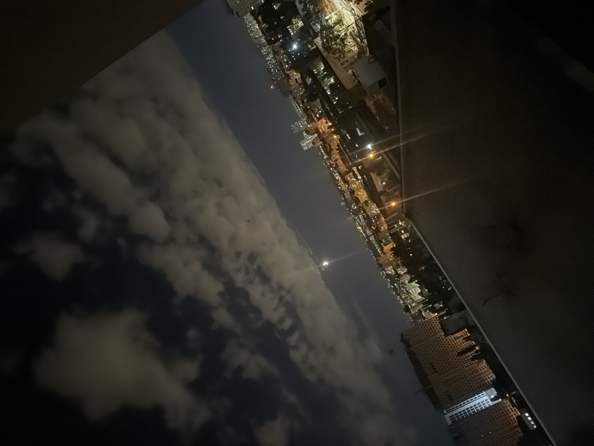 The image captures a nighttime view of a cityscape from a high vantage point. The sky is dark, with scattered clouds illuminated by an unseen light source, possibly the moon, which is visible as a faint crescent in the upper center of the frame. Below the sky, the city is spread out with numerous buildings, many of which are lit, creating a warm glow. The architecture appears to be modern, with taller apartment blocks and smaller residential or commercial structures. Lights are visible along streets, suggesting activity within the city. The perspective is angled, giving a sense of depth and vastness to the urban landscape. The foreground is dominated by a dark, silhouetted structure, possibly a balcony or rooftop, which frames the view of the city. The overall scene conveys a sense of urban life continuing into the night, with the city lights contrasting against the darkness of the sky. The presence of tall apartment buildings suggests a residential area within a larger urban environment. The image quality is slightly grainy, consistent with a low-light photograph taken at night.