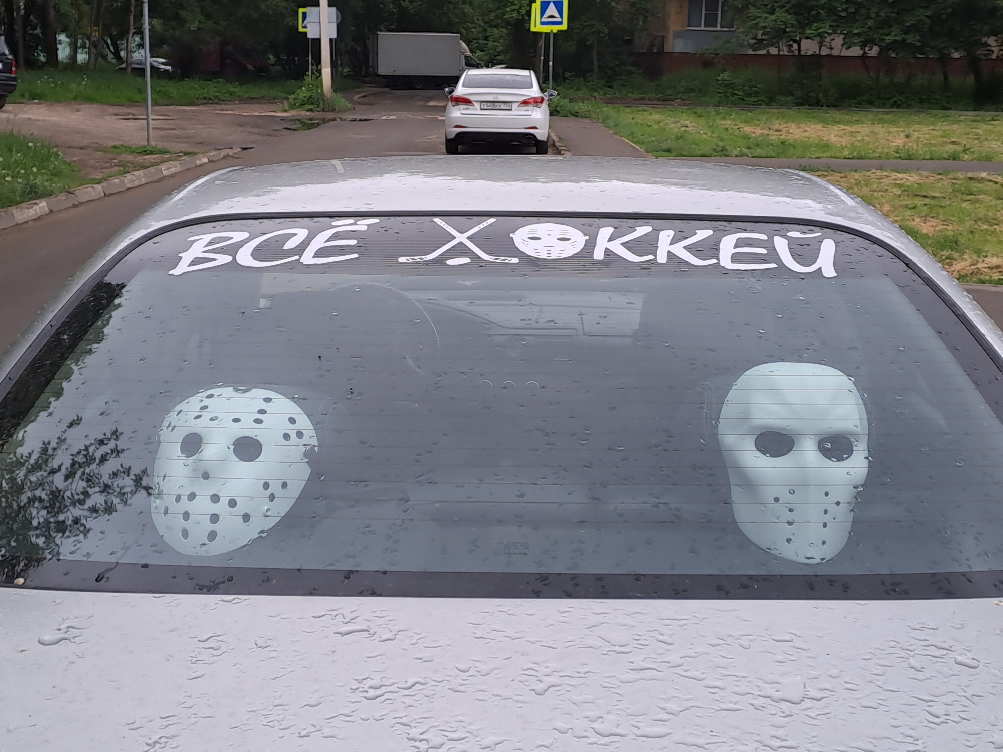 A silver car is parked in Elektrostal, Russia. The car's rear window is adorned with a white sticker that reads "ВСЁ ХОККЕИ" ("All Hockey"). Images of two hockey masks are placed inside on the rear window. Rain droplets cover the rear window and the car's surface. In the background, there is a street with trees and a white car parked in the distance. A pedestrian crossing sign and other traffic signs are also visible. A building is located to the right of the frame.