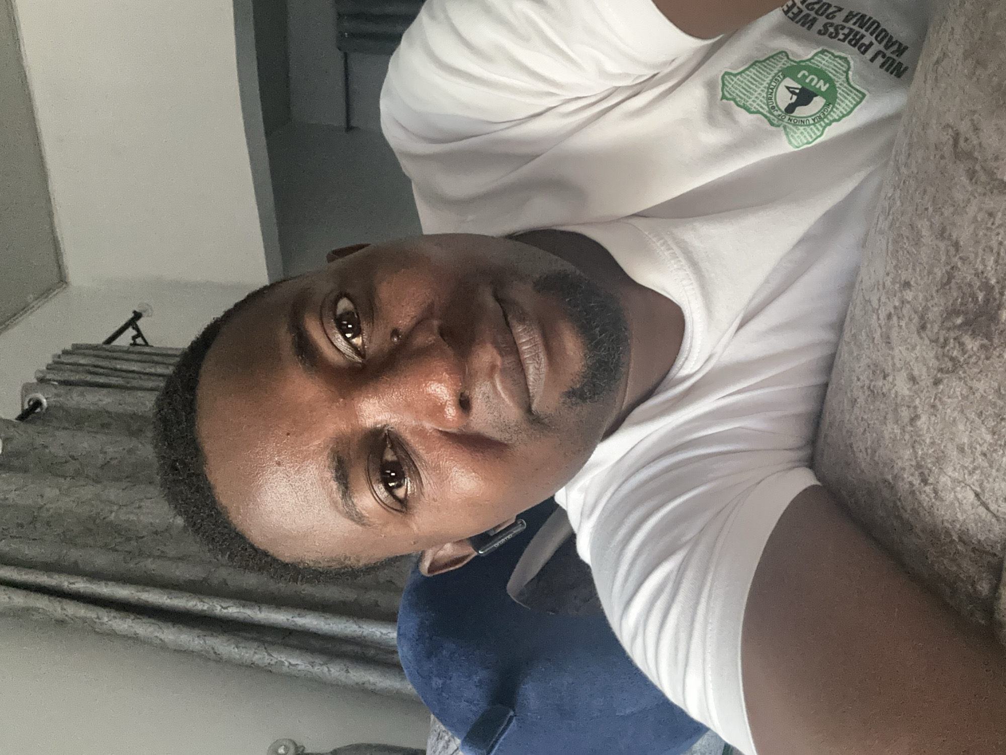 The image is a selfie taken by a Black man with dark, short hair and a goatee, wearing a white t-shirt. He is leaning on a textured, neutral-colored surface, possibly a couch or bed. To his left, a portion of a dark blue object, likely a pillow or cushion, is visible. In the background, grey curtains are drawn, and a portion of a wall and ceiling are visible. A logo on his t-shirt features a green outline of Nigeria with the text "NUJ" and "NIGERIA UNION OF JOURNALIST" within a circular emblem. Text below the emblem reads "NUJ PRESS WEEK KADUNA 2023". The man is looking towards the camera with a neutral expression. The scene appears to be indoors, in a residential setting, likely in Kaduna, Nigeria, given the text on his shirt.