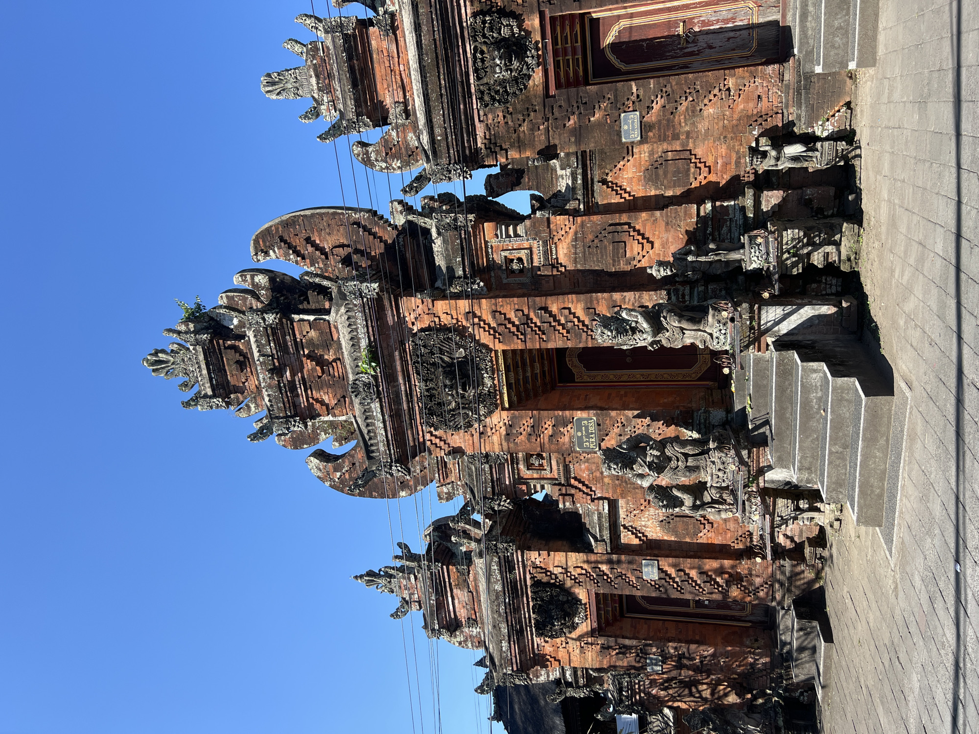The image shows an ornate brick temple gate in Sukawati, Indonesia. The temple gate is made of red brick and features intricate carvings and statues of mythical creatures and figures. Two large guardian statues stand on either side of the main entrance, which is reached by a set of stone steps. The temple has multiple levels and decorative elements, including layered roofs and finials. The sky above is clear and blue, indicating a sunny day. The foreground shows a paved walkway.