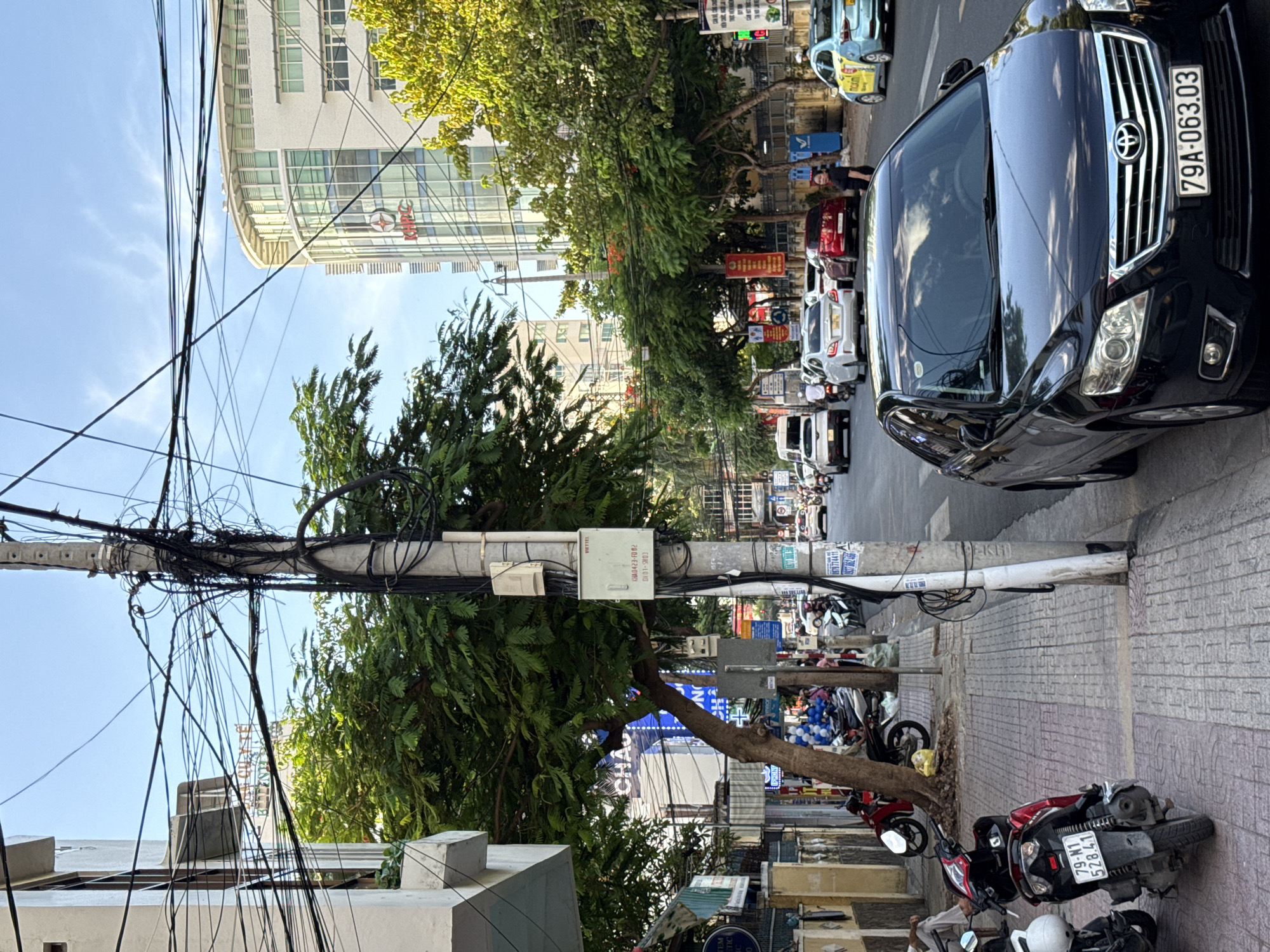 The image depicts a street scene in Nha Trang, Vietnam, characterized by a prominent utility pole covered in a dense network of electrical wires. In the foreground, a black Toyota Camry with license plate "79A-063.03" is parked along the side of the road. To the left, a sidewalk features parked motorcycles, including a red and black scooter with license plate "79-N1 528.41."
Across the street, traffic consists of cars and motorcycles. Several trees line the street, with lush green foliage. A modern, white, multi-story building with a rounded facade and visible windows is present in the background. The sky is partly cloudy. The overall impression is a busy urban street with a mix of modern architecture and everyday city life, punctuated by the visual clutter of numerous overhead wires.