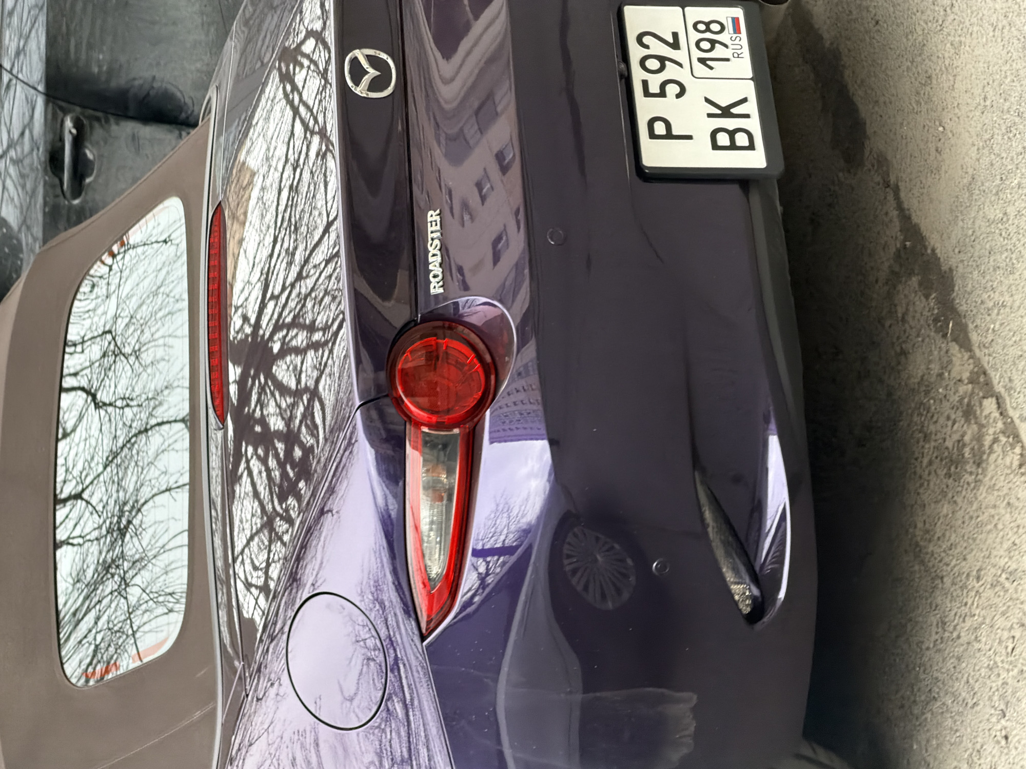 The media file presents a close-up, angled view of the rear left side of a dark purple Mazda MX-5 Miata Roadster, parked outdoors. The car features a beige soft top, partially visible on the left, and a prominent red taillight assembly.
Visible on the trunk are the Mazda logo and the word "ROADSTER" embossed below it. A Russian license plate is mounted on the rear, displaying the characters "P 592 BK 198 RUS". The "198" code indicates the Saint Petersburg region, aligning with the provided location context.
The car's reflective paint surface shows clear reflections of bare tree branches, suggesting a season without foliage, such as late autumn or winter. Reflections of a multi-story building with several windows are also discernible on the car's body, indicating an urban or suburban setting. The ground beneath the car appears to be damp concrete or asphalt.
The scene takes place during the daytime, likely under an overcast sky, given the diffused lighting and the absence of strong shadows. No people or active events are depicted; the focus is solely on the stationary vehicle. The car's fuel cap is also visible on the rear quarter panel.