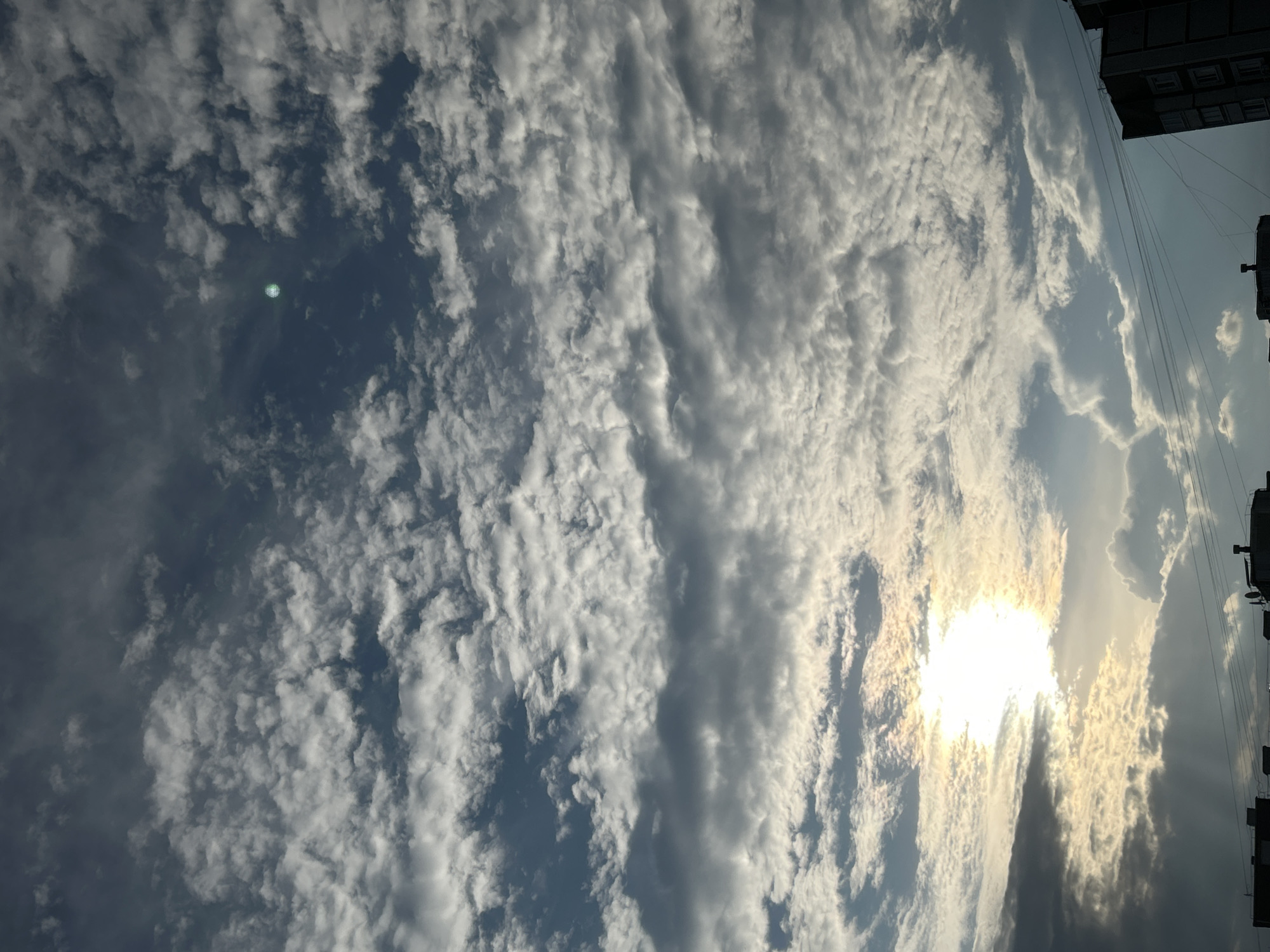 The image shows a sky with scattered clouds, illuminated by the sun which is partially obscured by clouds in the lower left portion of the frame. There is a bright green circular lens flare in the upper right quadrant of the sky. In the lower portion of the image, the tops of several buildings and utility wires are visible, suggesting an urban environment. The scene is likely Zelenograd, Russia, given the provided location context.