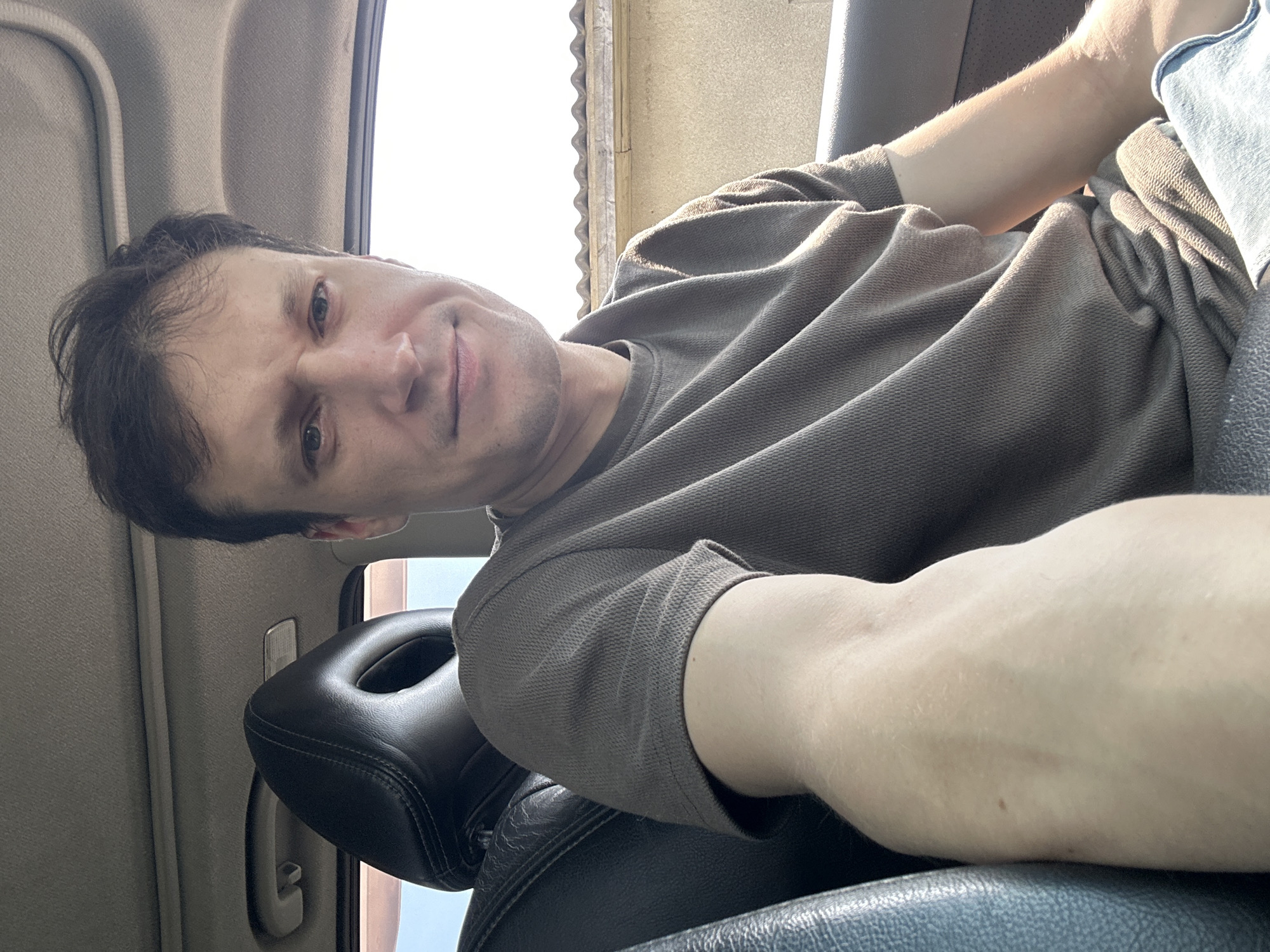 A man is sitting in the driver's seat of a car, facing forward and taking a selfie. He has short, dark hair and fair skin. He is wearing a brown t-shirt and blue jeans. The interior of the car is visible, including the black leather headrest and part of the dashboard. The window is open, and sunlight is entering the car. Outside, a tiled roof and a beige wall are visible. The scene is taking place in Shatava, Ukraine.