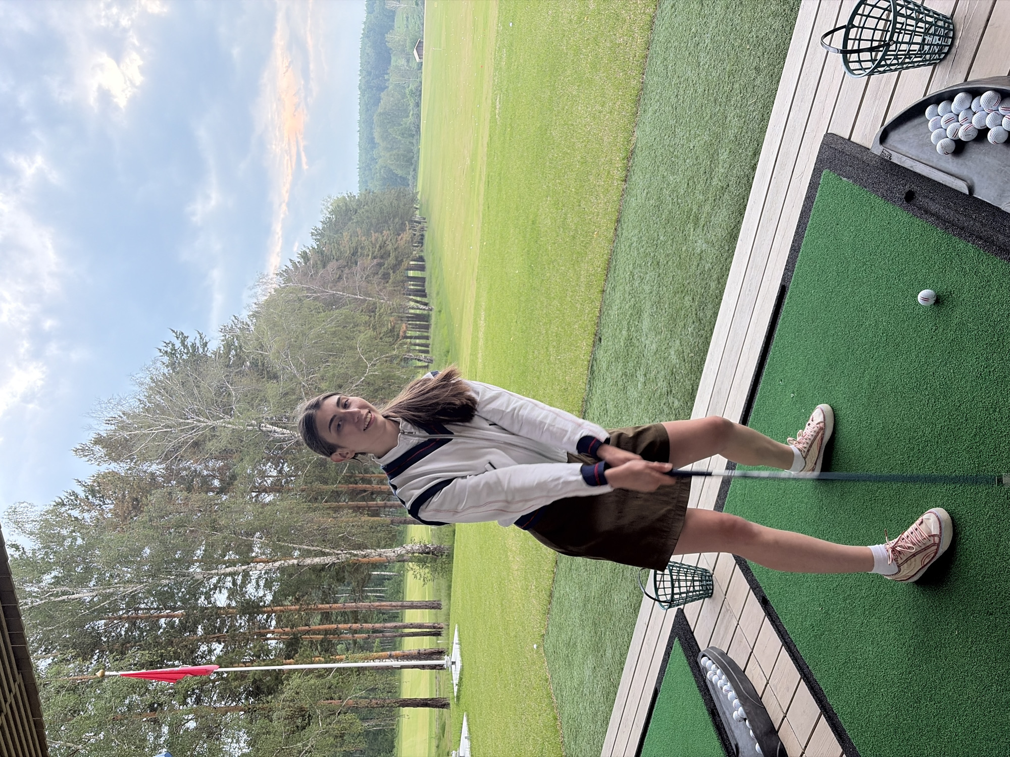 A young woman is pictured at a golf driving range, preparing to swing a golf club. She is wearing a white jacket with navy and red trim, a brown skirt, and pink sneakers. The driving range features artificial turf mats, a bucket of golf balls, and a large green field with trees in the background. The scene appears to be in an outdoor setting, possibly in the outskirts of Krasnoyarsk, Russia, given the prevalence of coniferous and deciduous trees typical of the region. The sky is partly cloudy with the sun setting in the distance, casting a soft glow over the landscape.