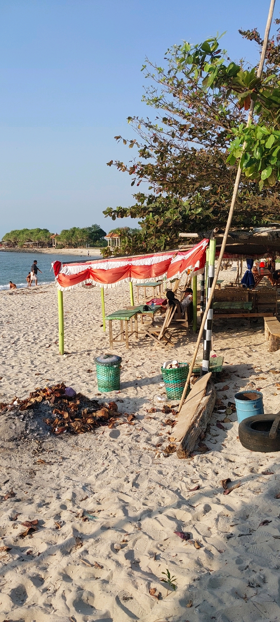 A photograph taken at a beach in Karanggondang, Indonesia, shows a serene scene with a focus on the sandy shore and the ocean. The sky is clear and blue, with a few scattered clouds. The sand is light-colored and appears to be clean, with no visible litter or debris. The ocean is calm and blue, with gentle waves lapping at the shore.
In the foreground, a covered seating area with red and white striped fabric shades the sand. Several tables and chairs are set up under the shade, some of which are occupied by individuals. To the left, a pile of dried leaves and debris lies on the sand, with two green woven baskets nearby. In the background, more trees and vegetation line the shore, and a few small buildings are visible on a distant island. A person is walking near the water's edge, and a few other individuals are seen in the distance. The overall impression is one of a peaceful and relaxing beach environment.
