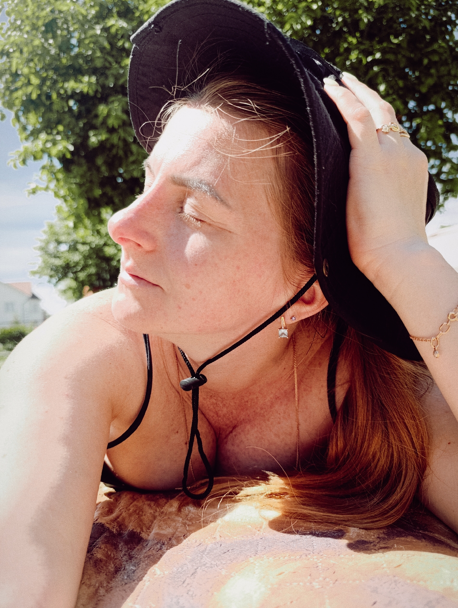 The image presents a close-up, slightly low-angle perspective of a female individual reclining outdoors under bright sunlight. The subject is depicted in profile, facing the left side of the frame, with her eyes closed. Her skin is fair, exhibiting freckles across her nose, cheeks, forehead, and upper chest/shoulder. She possesses reddish-brown hair, with some strands escaping from beneath a dark-colored, wide-brimmed hat. Her right hand, adorned with light-colored nail polish, rests on the side of her head, appearing to adjust or hold the hat. A gold-toned ring with multiple small decorative elements is visible on her right ring finger, and a gold-toned bracelet with linked circular components and a small drop charm is present on her right wrist. Her left ear displays two earrings: a small purple stud and a larger square-cut clear gemstone dangle earring. A black cord, secured with a black stopper, is visible around her neck and upper chest. She is wearing a black-strapped garment, consistent with a bikini top or a tank top. The individual is resting on a textured, multi-toned surface that appears earthy or patterned. The background features blurred green foliage of trees against a clear blue sky, with indistinct white residential buildings visible in the far distance. Strong direct sunlight illuminates the scene, casting pronounced highlights and shadows. The exact location in the city of Unknown, Unknown, cannot be determined from the visual evidence.