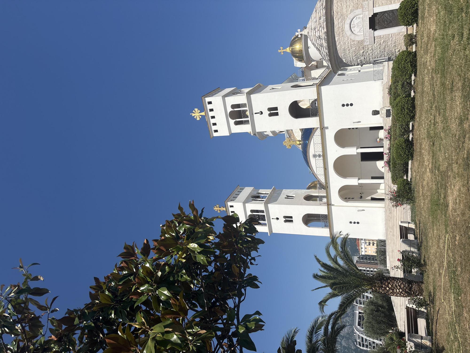 The image shows the Orthodox Church of St. John Vladimir in Bar, Montenegro. The church is white with two tall bell towers topped with gold crosses and domes. Lush trees and a palm tree are in the foreground. The sky is a clear, bright blue. Buildings can be seen in the background beyond the church grounds. The church is surrounded by grass.