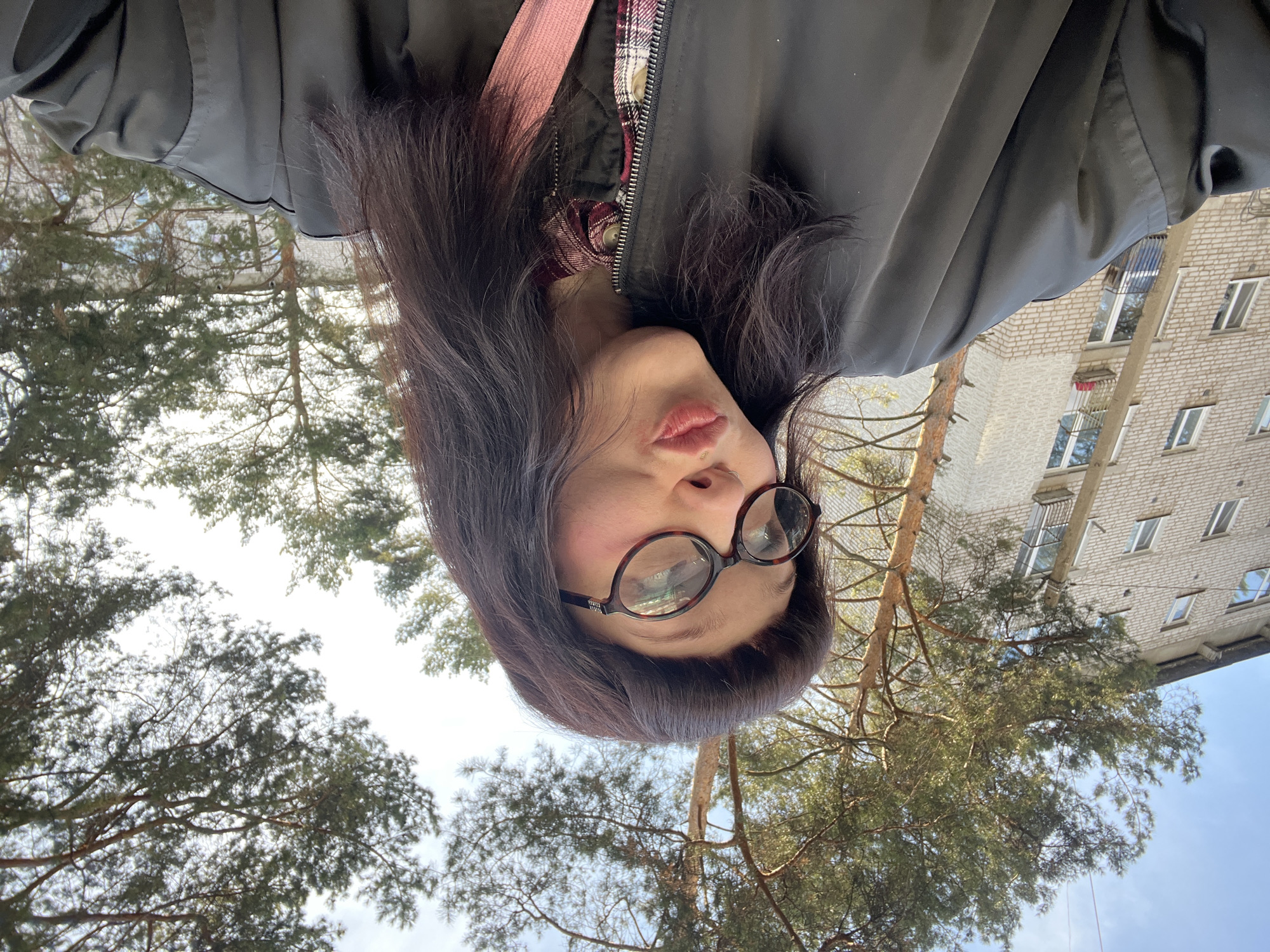 A selfie captures a woman in an outdoor setting. She has dark brown hair, wears glasses, and is dressed in a dark jacket with a pink strap visible across her torso. She is positioned in front of trees, and in the background is a multi-story brick building. The photograph appears to have been taken at an angle, with the camera pointing upwards. Based on the provided information, the location is Chaikovsky, Russia.