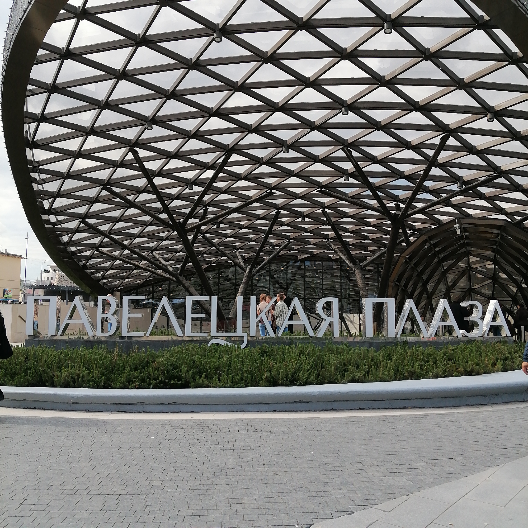 The image shows the entrance to the Paveletskaya Plaza shopping center in Moscow, Russia. The large, white Cyrillic letters spelling out "Paveletskaya Plaza" are positioned on a landscaped area with green bushes. Behind the sign is the shopping center's distinctive, modern architectural roof structure made of intersecting beams forming triangular patterns. Three people are standing in front of the letters, seemingly engaged in conversation. The foreground features a paved area leading up to the sign and the shopping center entrance. Parts of at least two people on the sides are visible.