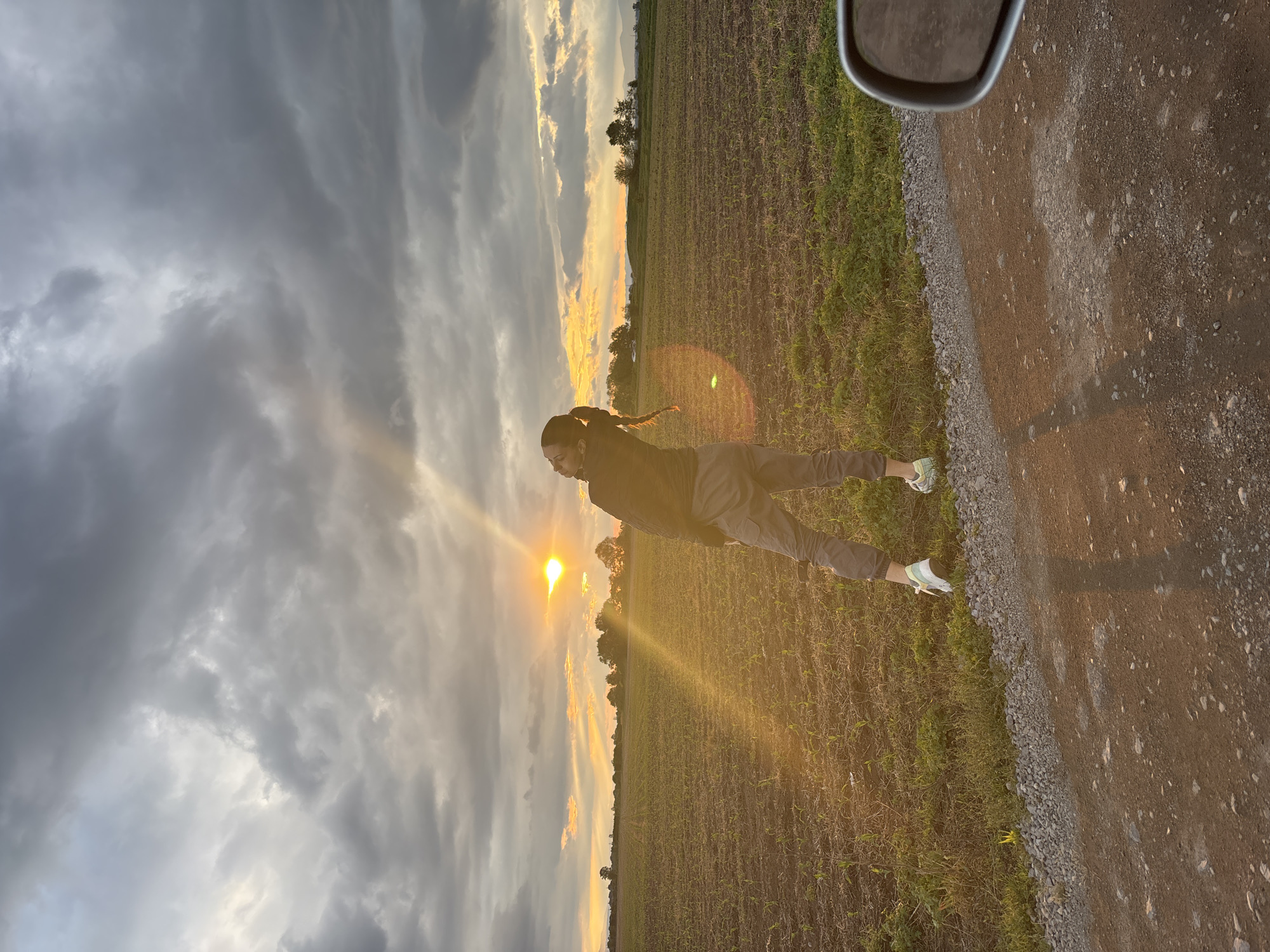 A person is standing in a field near a road, facing towards a sunset. The person has dark hair pulled back in a braid. They are wearing a dark jacket and light colored pants. The sun is setting on the horizon, creating a bright glow in the sky and reflecting off the ground. The field appears to be planted with crops. Part of a vehicle is visible in the upper right corner of the image. The environment suggests a rural or agricultural area. The location is in or near Doganovo, Bulgaria, as this was provided in the prompt.