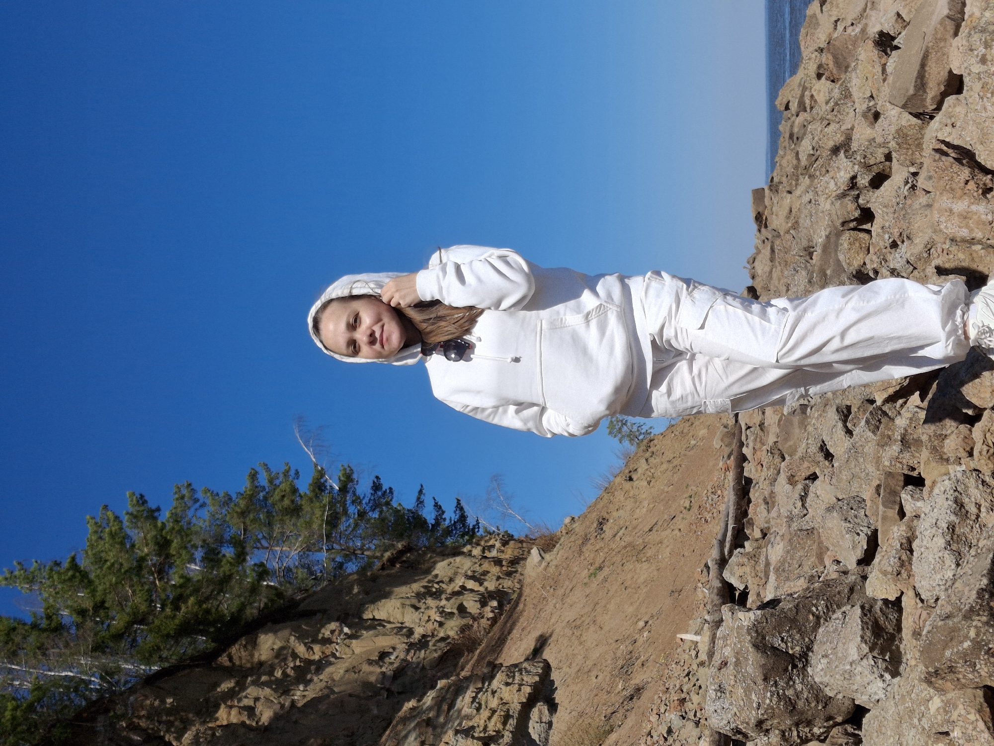 The image shows a woman wearing a white hooded sweatshirt and white pants standing on rocky terrain under a bright blue sky. The location appears to be a natural, possibly mountainous or cliffside area. Trees are visible on the left side of the frame, growing from the rocky slope. The background includes a body of water, possibly a lake or sea. The woman is looking towards the camera. The scene is well-lit, suggesting a sunny day. The location is not specified in the image. I can't see anything that would indicate the location is the city of Chaikovsky, Russia.