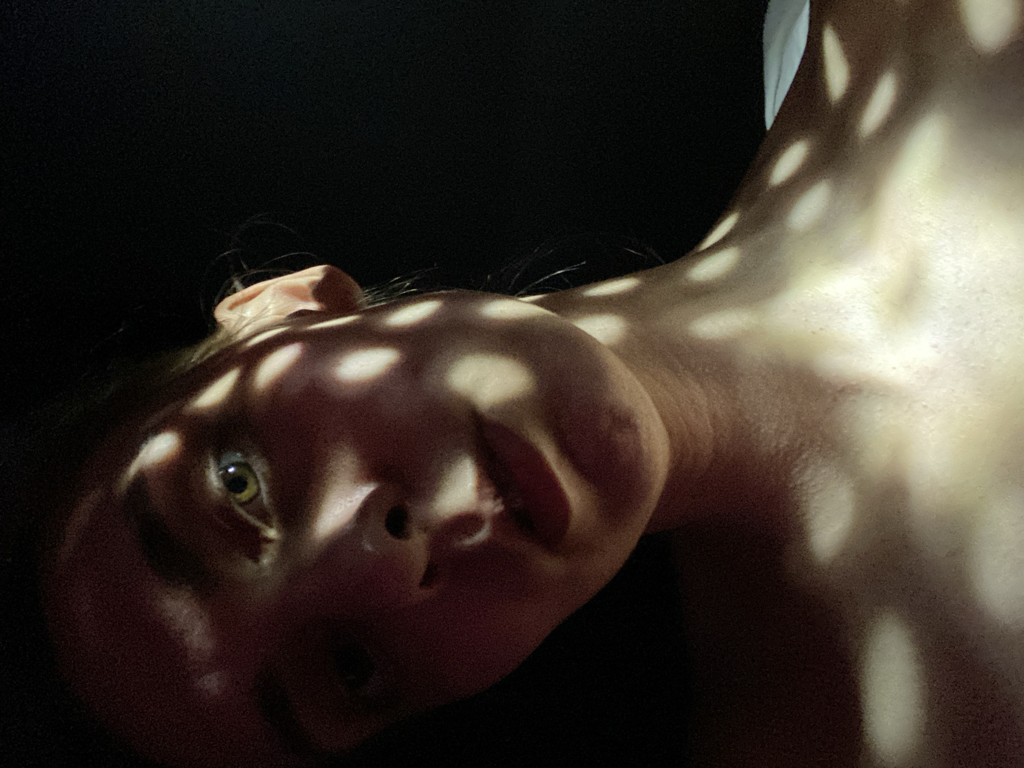 A close-up photograph features a person's face and neck, angled and illuminated with a pattern of dappled light and shadow. The person's face is turned towards the viewer, with one eye visible. The light creates a series of bright spots across the face and neck, contrasting with areas of deep shadow. The background appears to be dark and out of focus. There is no information to identify this photograph's location as the city of Chaikovsky, Russia.
