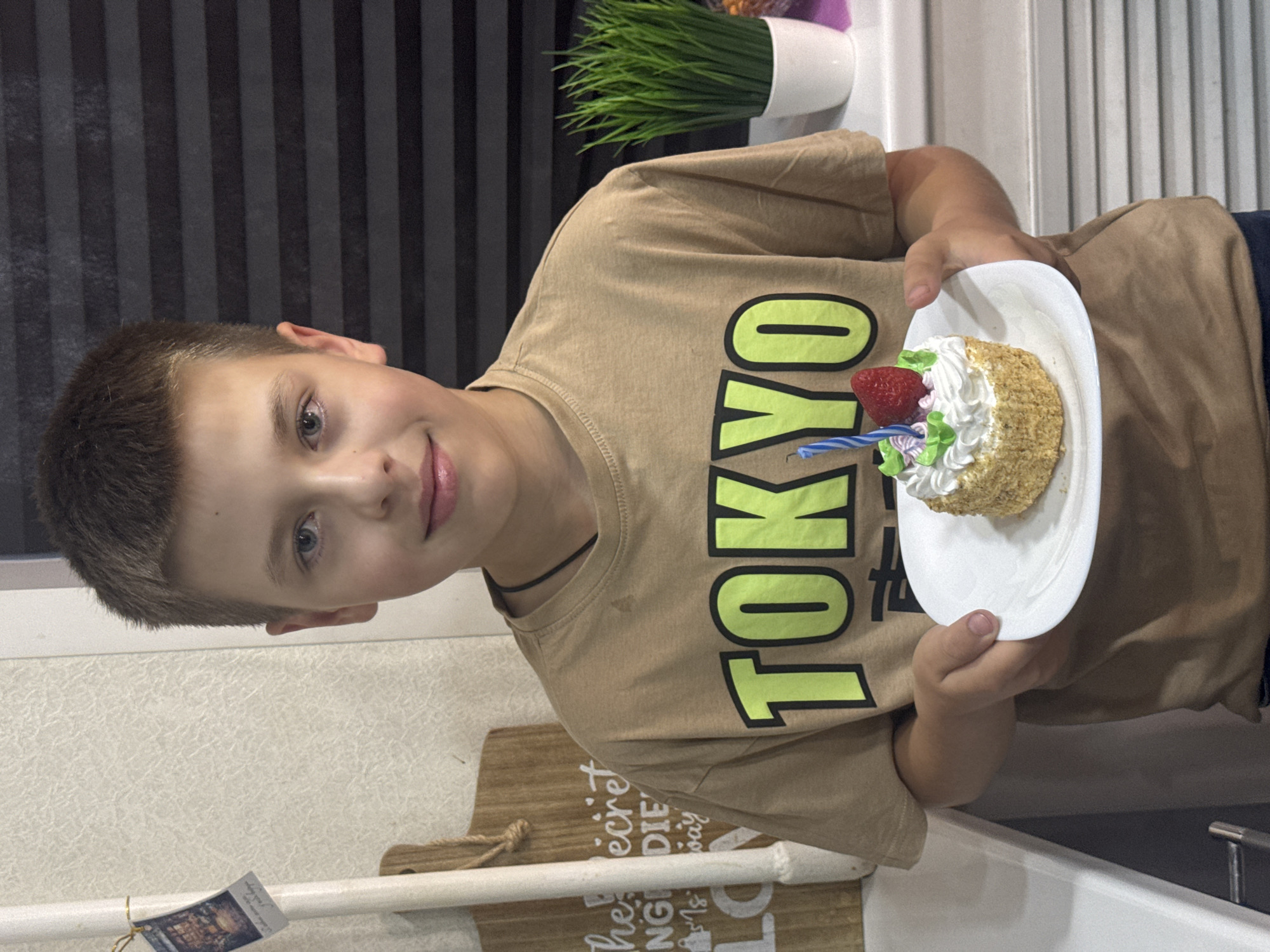 A young boy, likely under the age of ten, stands in what appears to be a kitchen or dining area, holding a small cake with a single lit candle and a strawberry on top. He is wearing a tan t-shirt with the word "TOKYO" printed in bright green letters with a black outline. The background features a textured wall, a wooden decorative board with text, and a plant in a white pot. The overall lighting suggests an indoor setting, possibly during the daytime. The boy's expression is neutral.