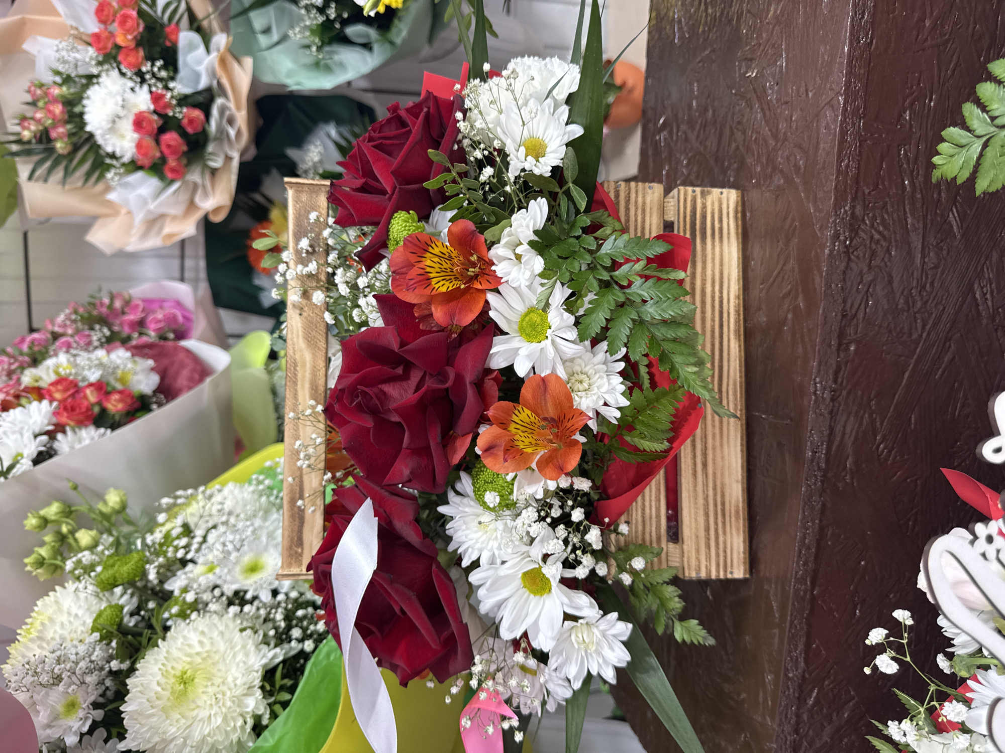 An indoor close-up shot displays a floral arrangement in a wooden crate. The arrangement features several deep red roses as the focal point, surrounded by white daisies, orange alstroemeria, and a sprig of green foliage. A white ribbon is tied around the arrangement, with a pink ribbon visible on the left side. The wooden crate is filled with red paper lining. In the background, other floral arrangements are visible, suggesting a flower shop or market setting. The surface beneath the arrangement is a dark brown, textured material. The location is identified as a flower shop in Kamianske, Ukraine.