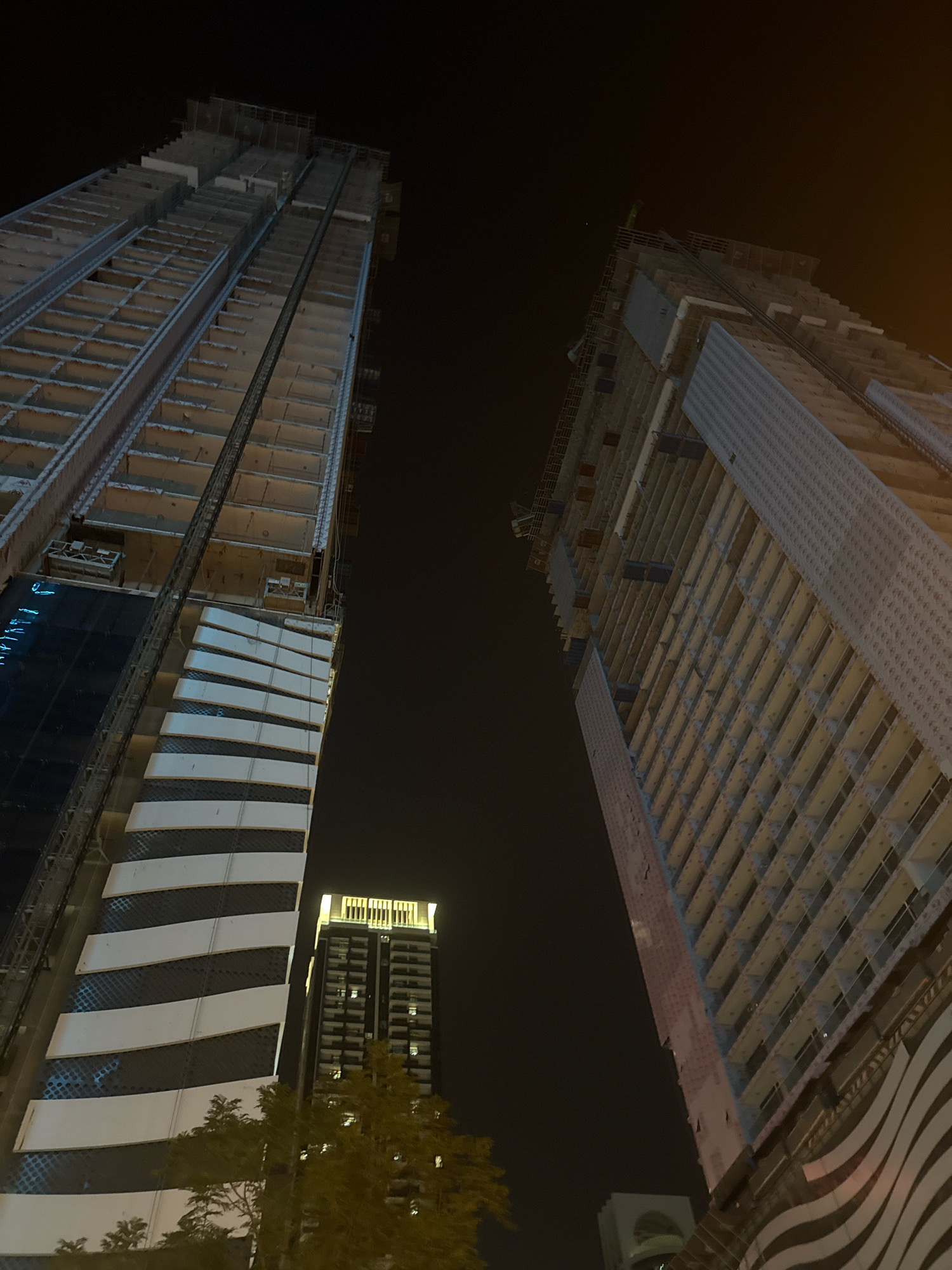 This image depicts a nighttime view of two under-construction skyscrapers in Dubai, United Arab Emirates. The perspective is from below, looking upwards at the imposing structures. The building on the left, partially completed, features a facade with alternating horizontal white bands and dark sections, possibly glass. The building on the right is in an earlier stage of construction, with its skeletal framework and concrete floors visible. In the background, a shorter, illuminated building stands complete. The sky is dark, suggesting it is night. Trees are visible in the foreground, partially obscuring the lower sections of the buildings. The overall scene conveys a sense of ongoing development and large-scale urban construction characteristic of Dubai.