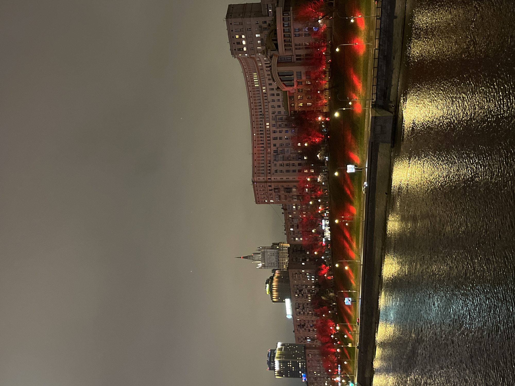 The image depicts a nighttime cityscape with several buildings, streets, and a body of water, likely a river. The buildings vary in height and architecture, some appearing older and more ornate, while others are modern. They are illuminated with artificial lights, some with a reddish hue along the streets. The water reflects the light from the city, creating a shimmering effect. The sky is overcast or foggy, giving the scene a muted, atmospheric quality. The location appears to be an urban area with waterfront development.
