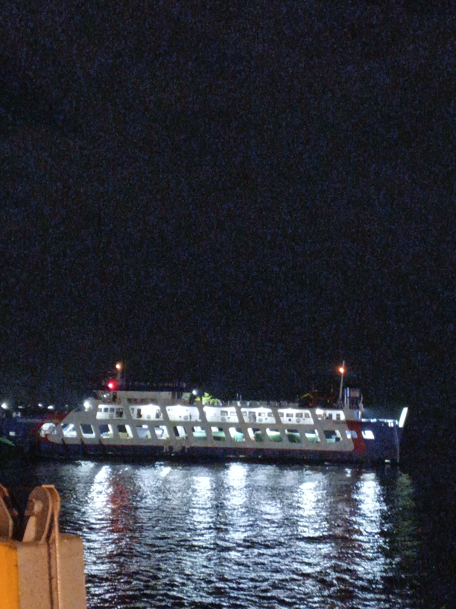 An exterior, wide shot captures a ferry at night on the water in Banyuwangi, Indonesia. The ferry is lit up with interior lights showing through the windows. It appears to be a large passenger ferry with multiple decks. The water is dark and reflects the ferry's lights, creating streaks of light on the surface. In the foreground, part of a dock or pier is visible. The sky is dark, indicating it is nighttime.
