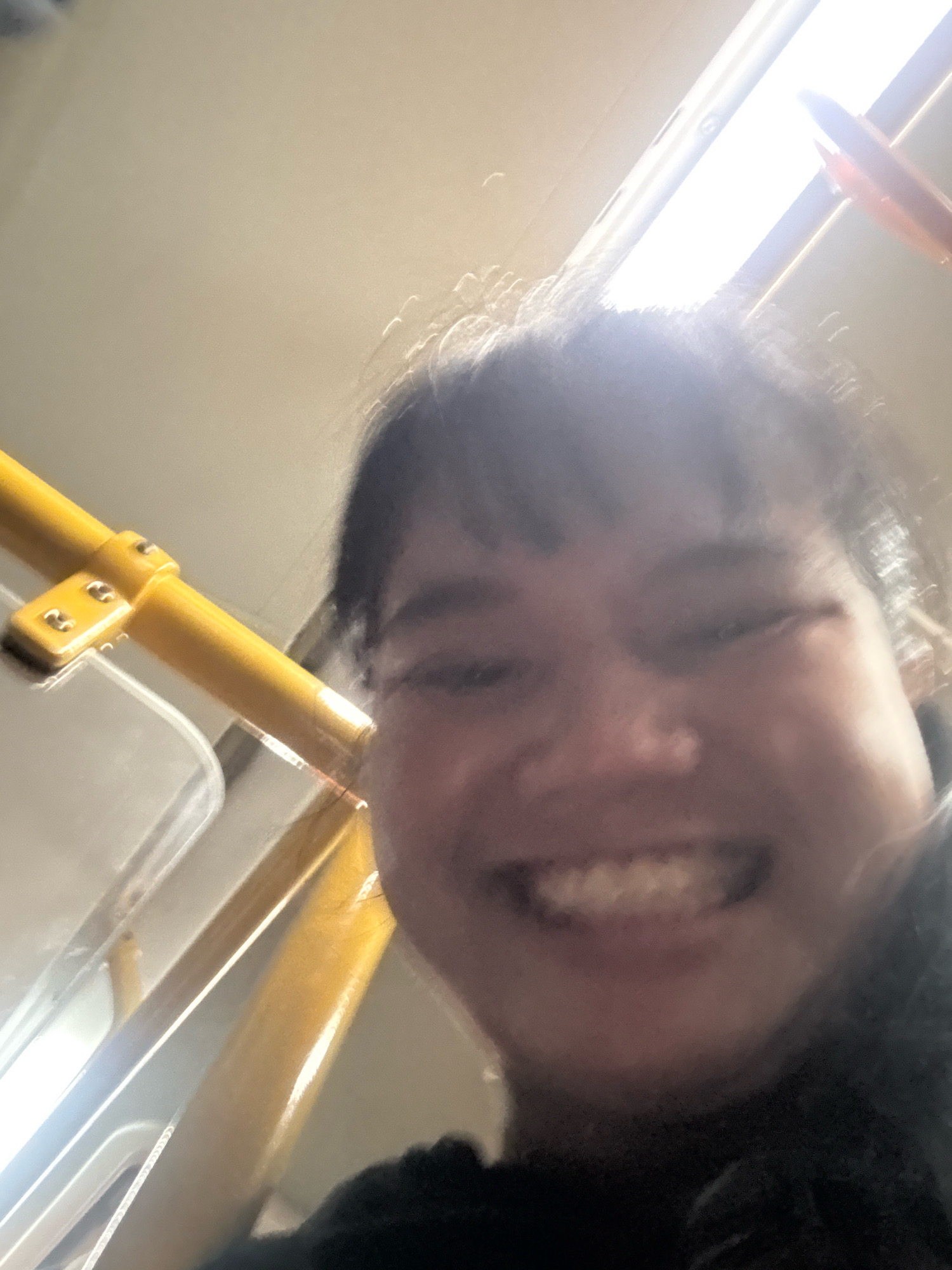 A selfie taken from a low angle captures a person's face with a bright light source behind them, creating a halo effect. The person is smiling, with their teeth visible. They appear to be on a form of public transportation, as indicated by the yellow handrails and a grey seat visible in the foreground. The environment is lit by overhead fluorescent lights. The image is blurry and lacks specific identifying features of North Jakarta, Indonesia.