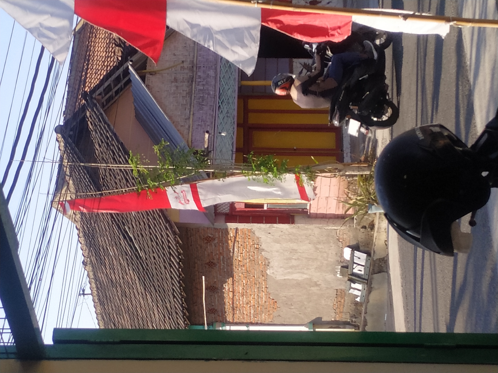 The image shows a street scene in Batan, Indonesia, featuring a brick building with a tiled roof and a motorcycle rider. The building has a weathered facade with exposed brickwork and a section of grey plaster. A red and white Indonesian flag hangs from a bamboo pole in front of the building, partially obscuring a smaller flag. Behind the building, there's another structure with a colorful facade, featuring yellow and red doors and purple siding. The rider, wearing a helmet, is on a black motorcycle and appears to be moving away from the camera. In the foreground, a black motorcycle helmet is visible. The environment suggests a sunny day with clear blue skies and overhead power lines.