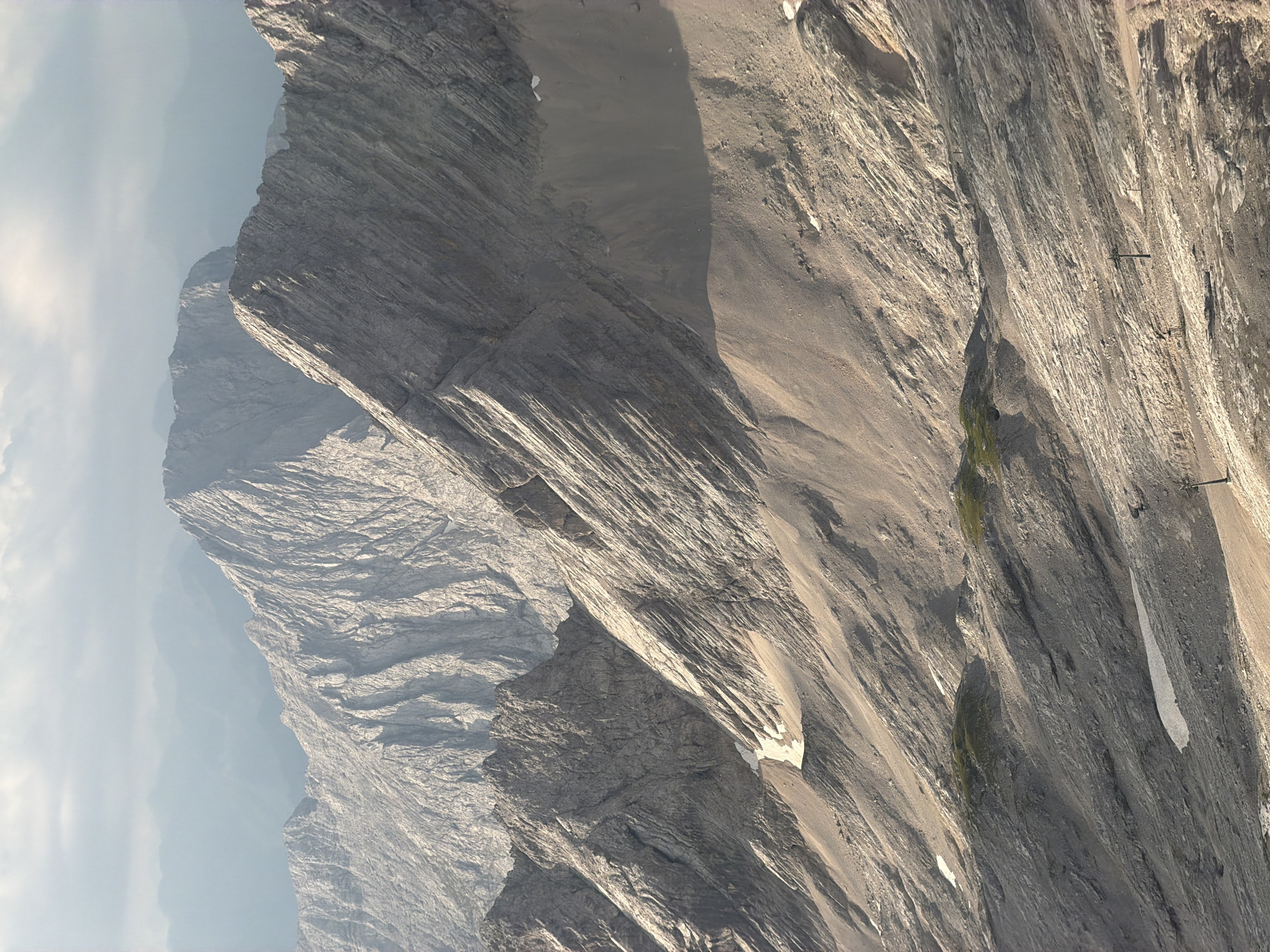 This image captures a scenic mountain landscape, likely taken from a high vantage point in the Bavarian Alps, near Garmisch-Partenkirchen, Germany. The scene is dominated by rugged, gray, rocky mountain peaks, with layered striations evident on their surfaces. Patches of sparse green vegetation are visible in some areas, interspersed with what appears to be snow or ice. The atmosphere is hazy, with distant mountain ranges fading into a light blue mist. The foreground features a steep, rocky slope with some scattered debris. Several dark, vertical structures, possibly ski lift pylons or weather stations, are present in the lower section of the image. The lighting suggests it is either early morning or late afternoon, with sunlight casting long shadows and highlighting the textures of the mountains.