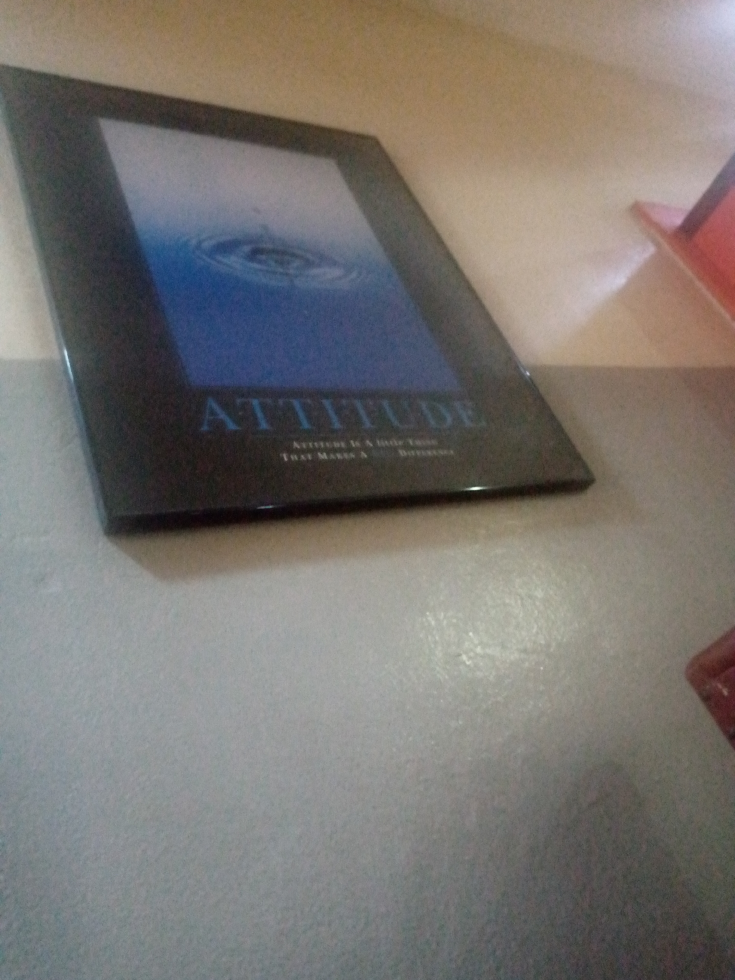 The image shows a framed poster leaning against a wall or other surface in a location of Unknown, Unknown. The poster has a black frame and features a blue image of ripples in water. The word "ATTITUDE" is prominently displayed in blue font on the poster. The surrounding area appears to be a room with a neutral-colored wall. A red object is visible in the upper-right corner of the image.