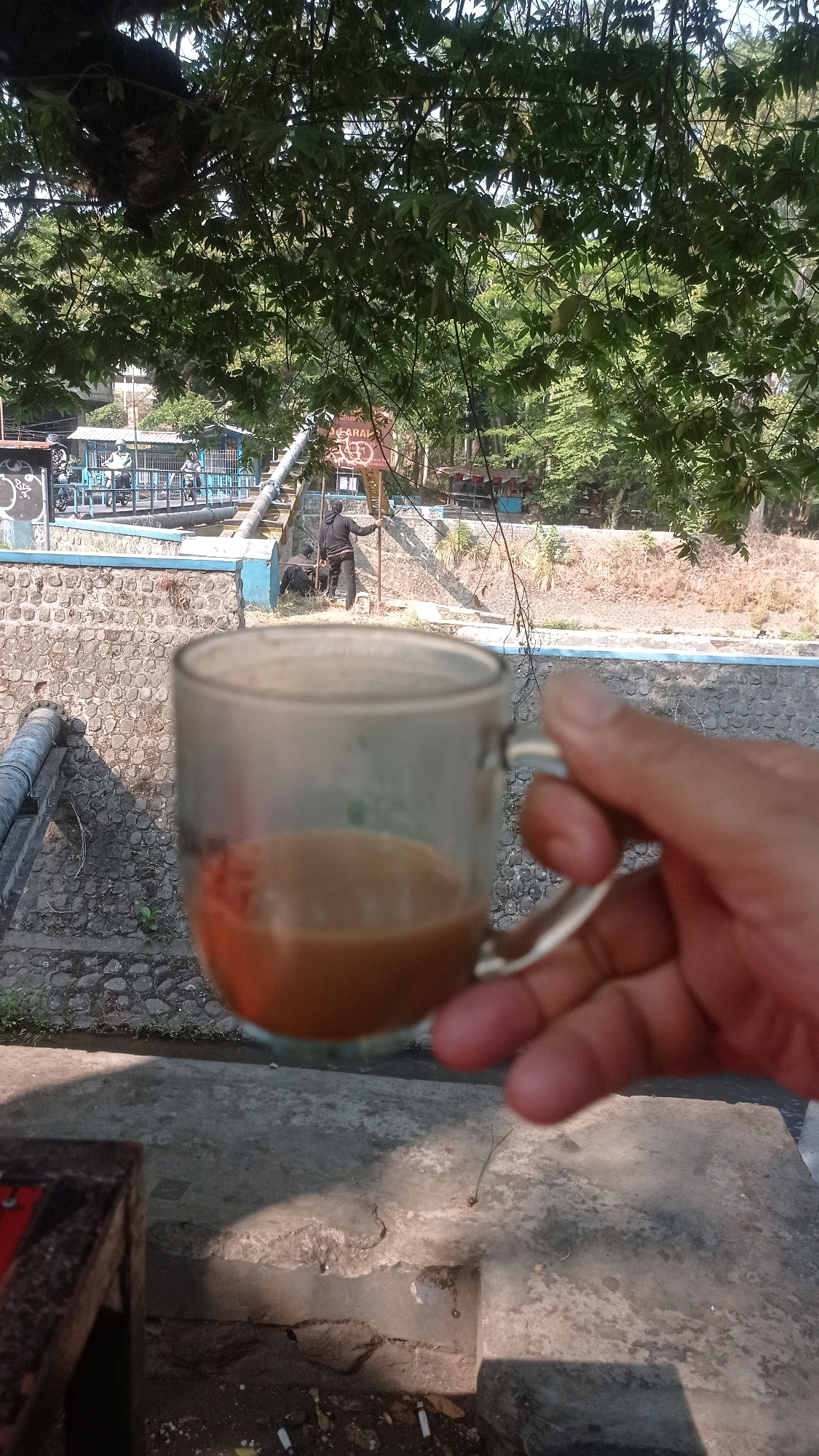 A hand holds a clear glass mug filled with a light brown beverage, likely coffee or tea, in the foreground. The mug is positioned to the right of the frame, partially obscuring the background. The background reveals an outdoor scene with a stone-lined channel or embankment. On the embankment, two individuals are present. One, wearing dark clothing, stands and appears to be interacting with a pole or support. Another person is seated near a dark object. A blue railing runs along the top of the embankment. Beyond the embankment, there is a bridge with several people on motorcycles. Trees with green foliage overhang the scene from the top of the frame. The ground in the immediate foreground is a rough, grey concrete surface with some debris, including what appear to be cigarette butts. The overall impression is a relaxed outdoor setting, possibly a park or a riverside area, in a place like Mulyoagung, Indonesia.