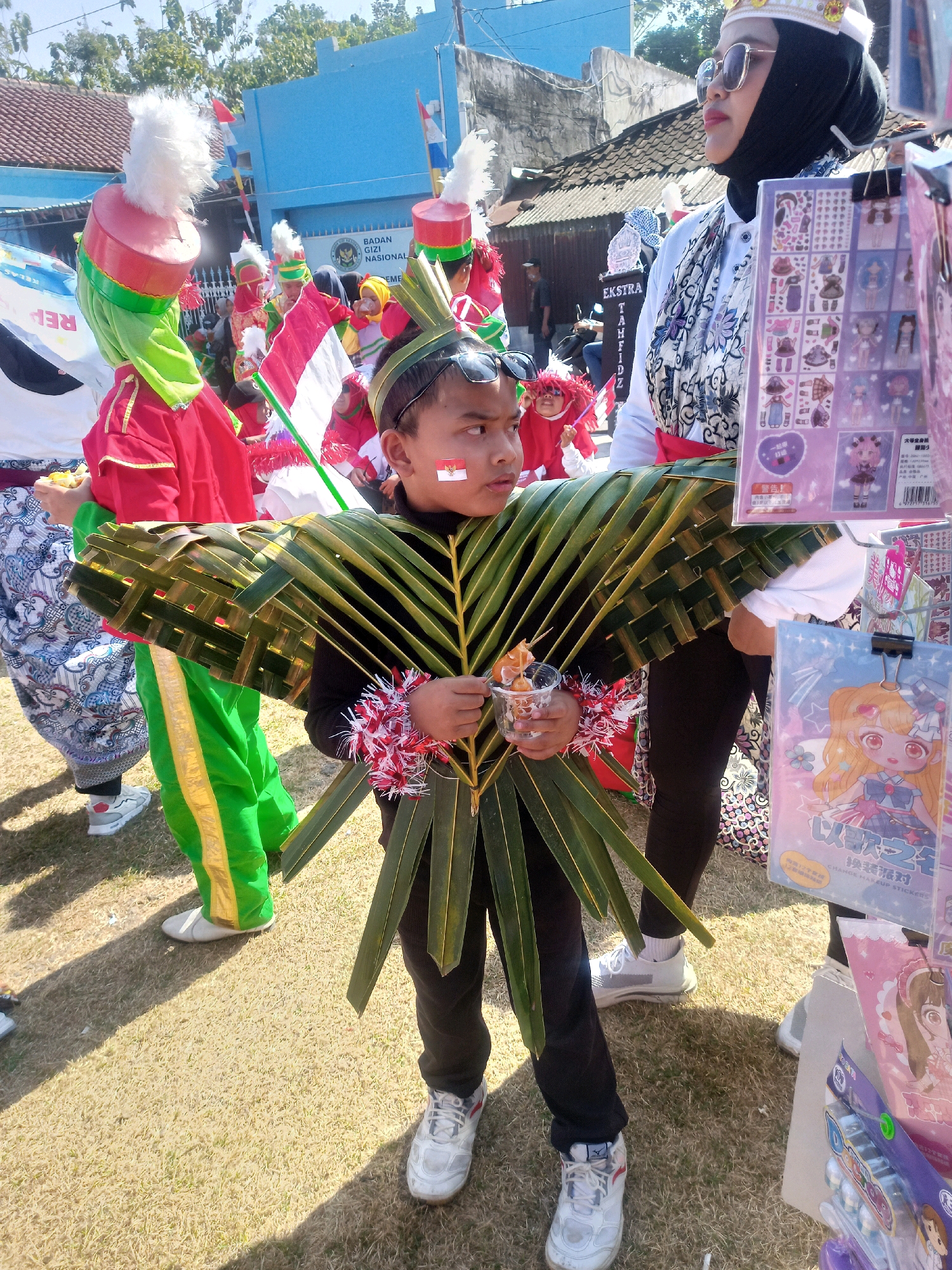The image depicts a street scene in Sumberlawang, Indonesia, likely during a parade or festival. In the foreground, a young boy is dressed in a unique costume made of palm leaves, forming a fan-like structure around him. He wears black pants and white sneakers. On his cheek, a small Indonesian flag is painted. He is holding a clear plastic cup containing small food items.
Behind the boy, other participants are visible, some in elaborate costumes. To the left, individuals in red and green traditional attire with tall, feathered hats are present, some holding Indonesian flags. To the right, a woman wearing a black hijab, white shirt, and a patterned scarf is standing, looking to the side. In front of her, a display of stickers featuring cartoon characters is set up.
The background shows a blue building with signage that appears to be for "BADAN GIZI NASIONAL". The environment is outdoors, with dry grass on the ground and trees and buildings visible in the distance under a clear sky. The overall impression is of a community event with a focus on cultural representation and festivity.