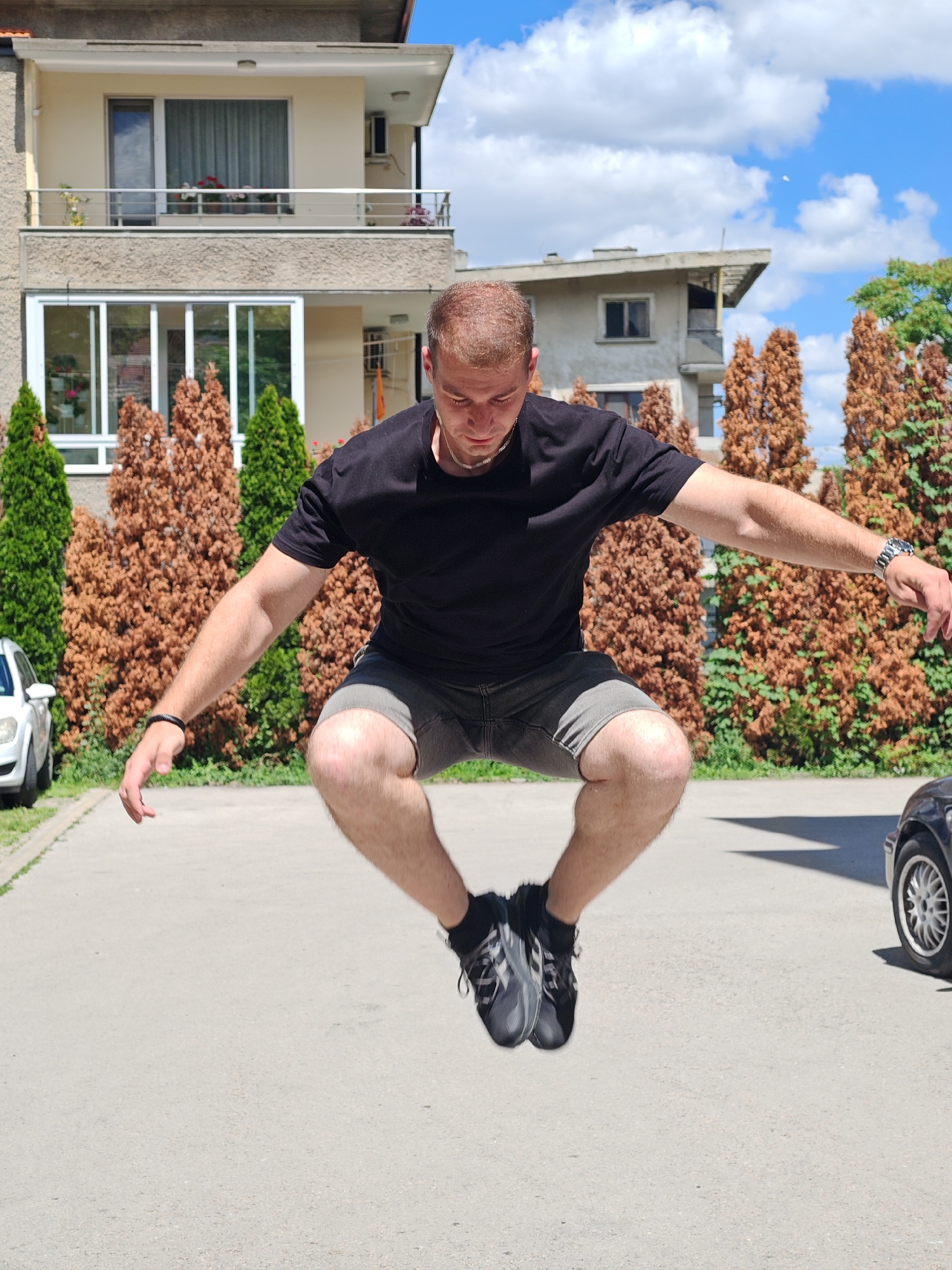 A young man with short, light brown hair is captured mid-air, jumping in a paved outdoor area. He is wearing a black t-shirt, grey shorts, and athletic shoes. His arms are extended to the sides, and his legs are bent at the knees. Behind him, a two-story residential building with a balcony and a smaller building with a slanted roof are visible, along with several rows of evergreen and brown, dried bushes. A white car is parked on the left, and the rear of a black car is on the right. The sky is blue with scattered clouds. This scene is located in the city of Shumen, Bulgaria.