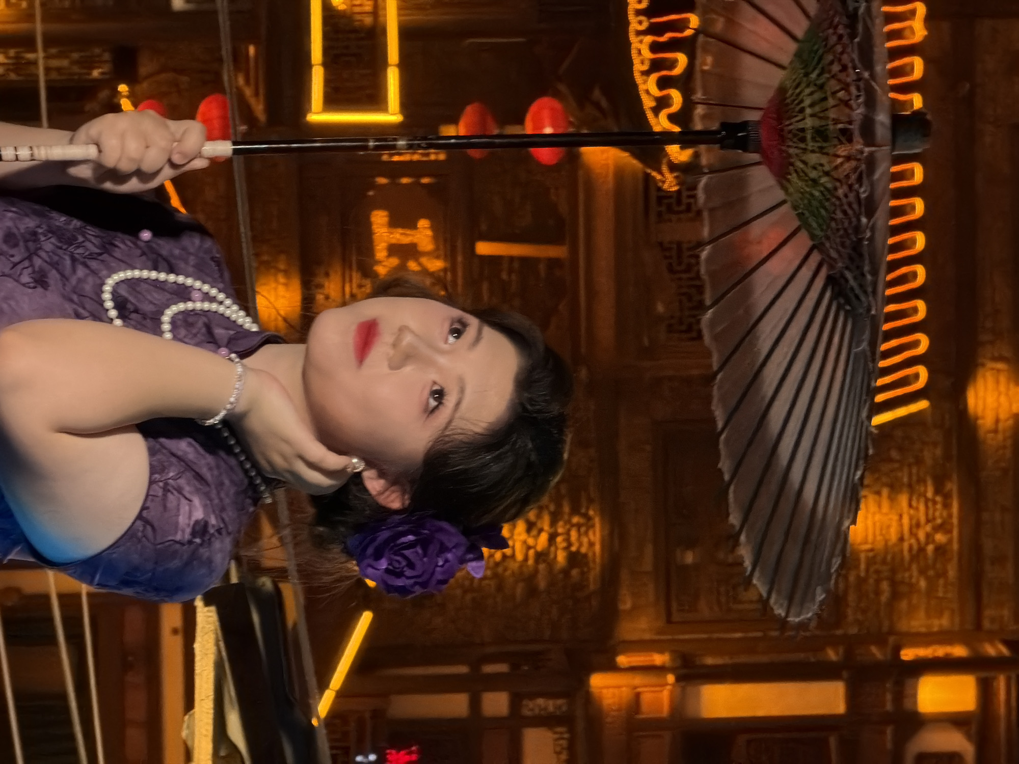 In the city of Yuzhong District, China, a young East Asian woman stands in front of an ornate wooden background illuminated by warm, orange lighting. She is dressed in a traditional purple qipao with a floral pattern and wears a multi-strand pearl necklace and a silver bracelet. A large, decorative purple flower is pinned to her hair. She holds a dark umbrella with a patterned canopy, resting its handle on the ground. Her head is tilted slightly upwards as she looks off to the side, her right hand touching her jawline. Red lanterns are visible in the background, as well as neon lights that accent the architectural details of the environment.