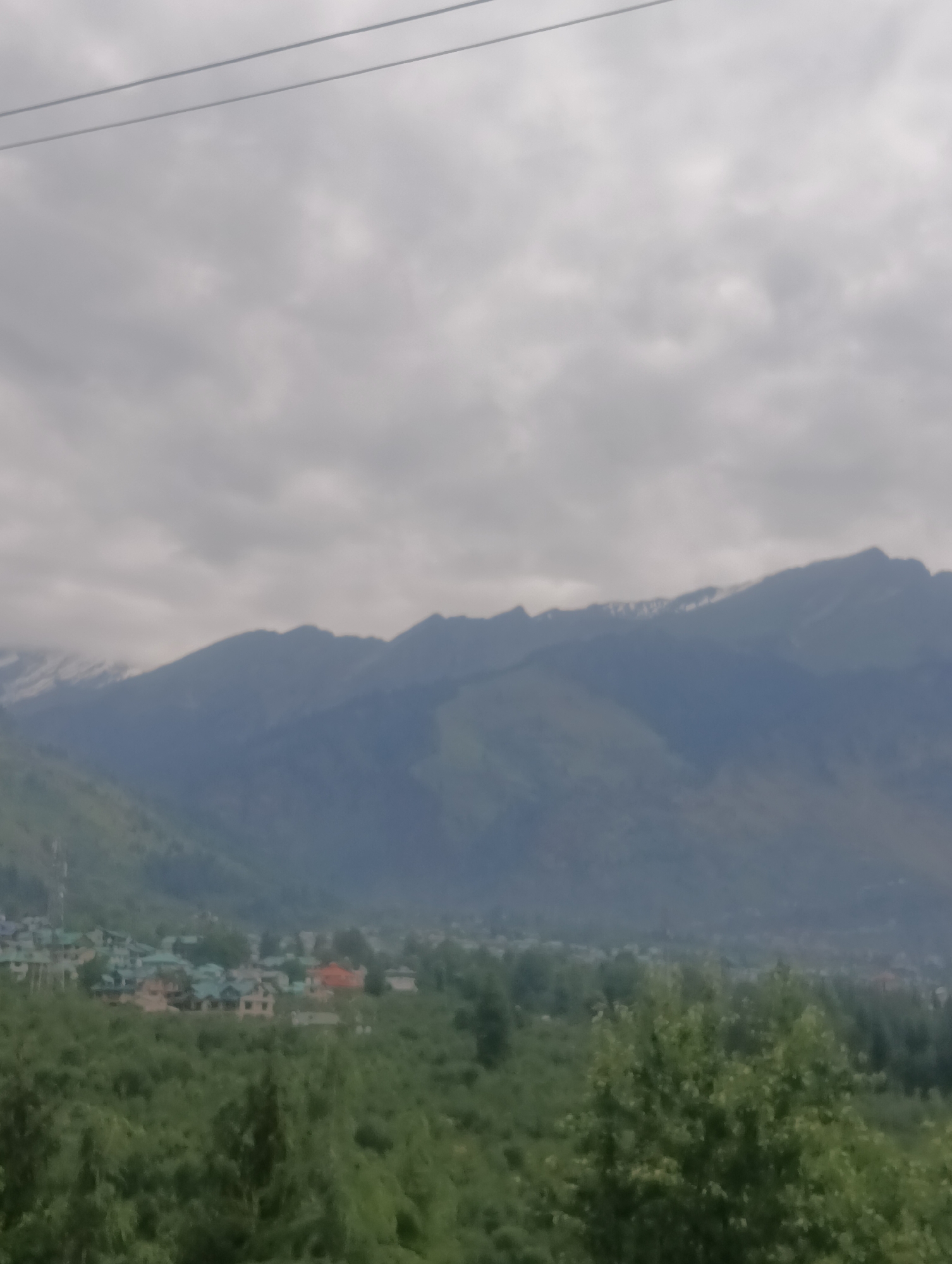 The image depicts a hazy mountain landscape in Shanag, India. In the foreground, dense green foliage obscures the lower portion of the scene. In the mid-ground, a cluster of buildings with predominantly green roofs are visible, suggesting a small settlement or village. Beyond the buildings, rolling hills rise, covered in a lighter shade of green vegetation. The background is dominated by a series of imposing mountains, their peaks partially obscured by clouds and mist, with some snow visible on the higher slopes. The sky is overcast, with a thick layer of gray clouds that diffuse the light, contributing to the overall hazy atmosphere. Two thin, dark horizontal lines are visible in the upper portion of the image, likely power lines. There are no people or animals clearly discernible in the image.