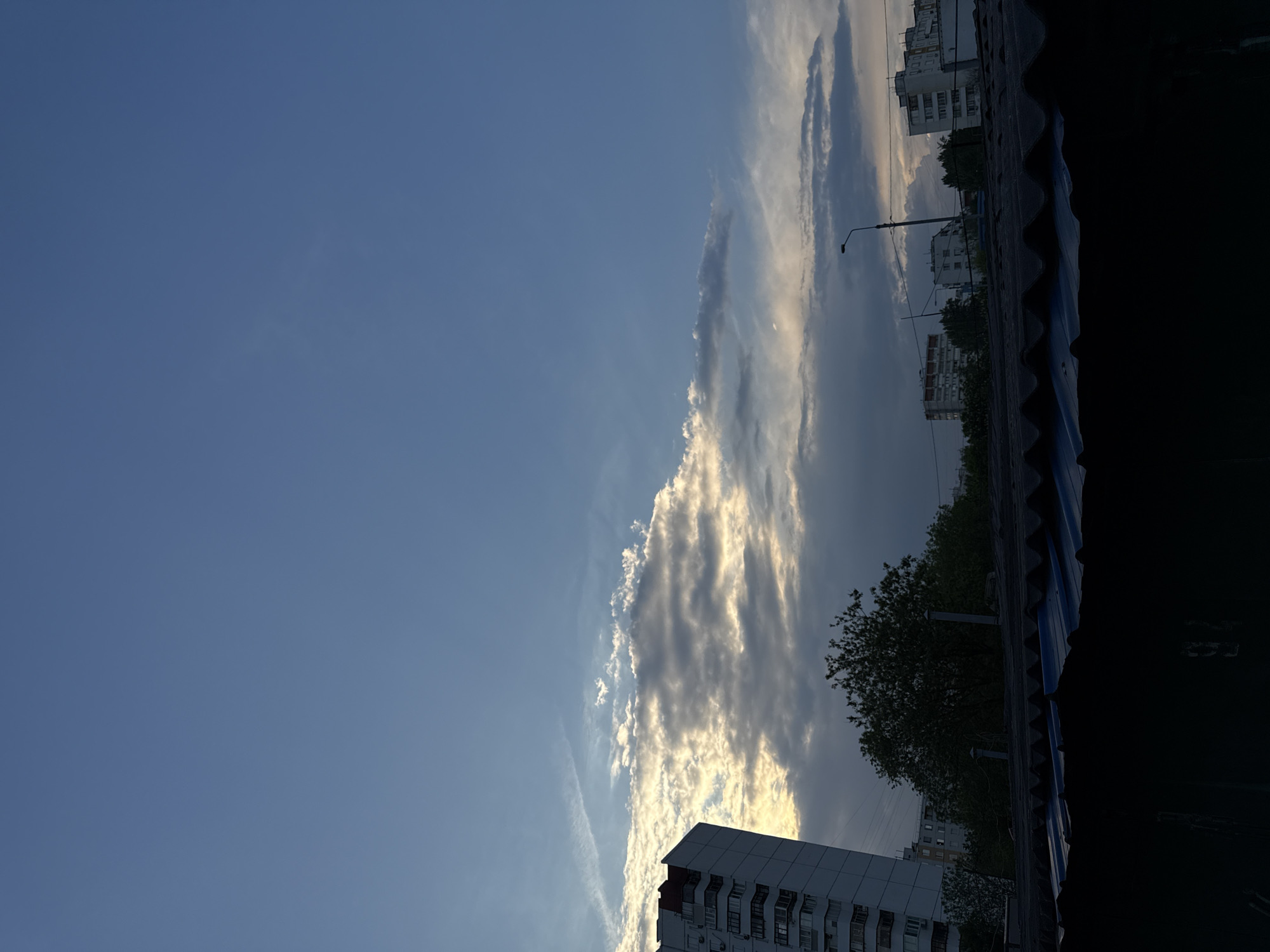 The image shows a view from what appears to be a high vantage point, possibly a roof or upper floor of a building, in an urban area. The location is likely Moscow, Russia, suggested by the architectural style of the residential buildings in the distance.
The focus is on the sky, which is predominantly blue with elongated, layered clouds catching the sunlight, giving them a bright, highlighted appearance.
In the foreground, parts of buildings are visible. To the bottom left, a corner of a high-rise building is seen. Towards the top right, the edge of a roof, potentially the one the photo was taken from, is visible, with dark, corrugated material. Behind it, there are residential buildings, trees, and a lamppost.
The scene conveys an early evening or late afternoon atmosphere due to the light and shadows. There is no human presence visible. The image captures a contrast between the natural beauty of the sky and the urban environment below.