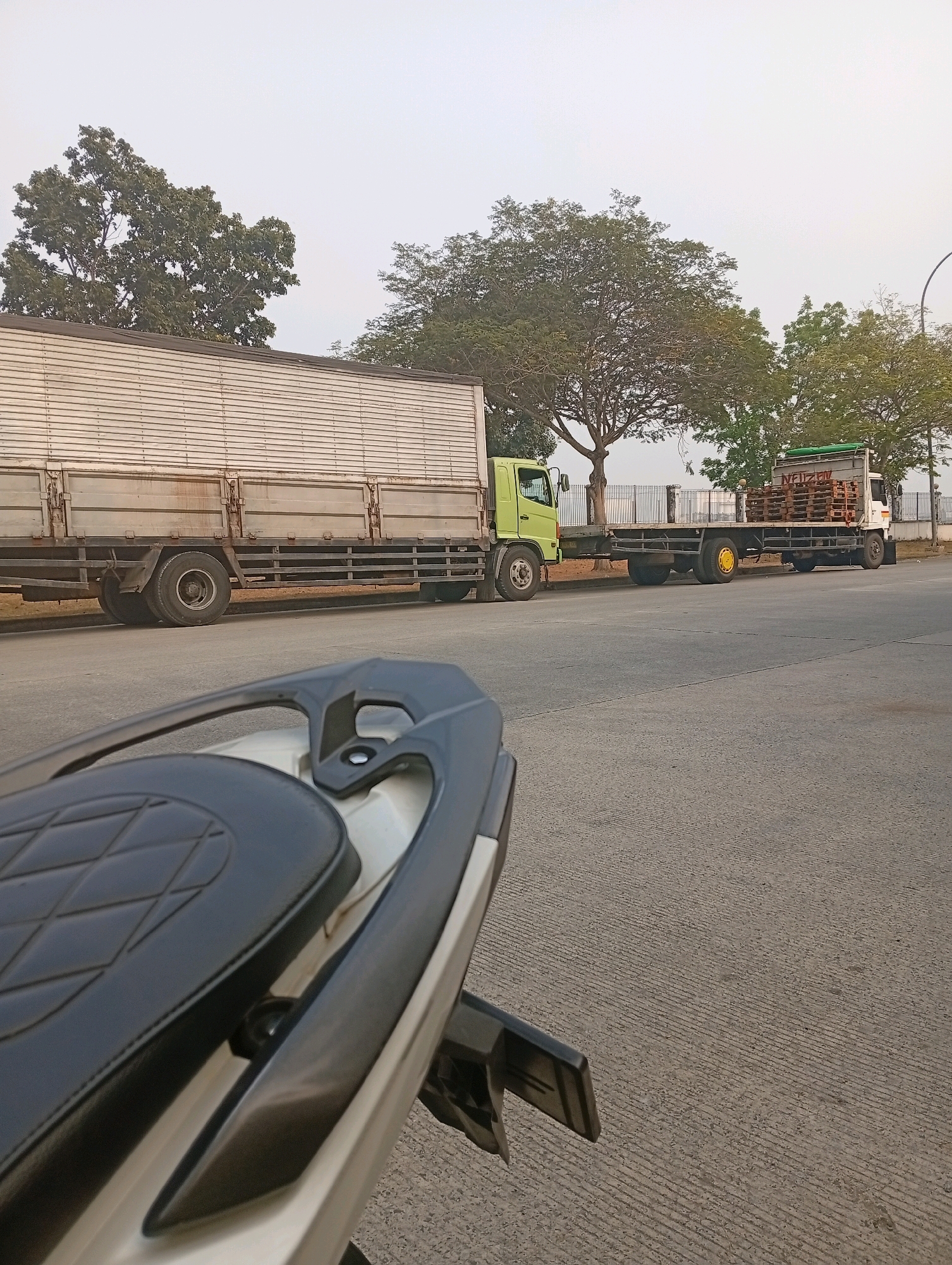 A light-colored box truck and a flatbed truck with wooden pallets are parked on a paved road in Sukamahi, Indonesia. The box truck, mostly obscured by a motorcycle seat in the foreground, is light green and white. The flatbed truck, white with a green top, has a white sign on its side. Trees with lush green foliage line the road in the background, behind a low fence. The sky is overcast and hazy. The foreground shows the rear of a motorcycle, with its black diamond-patterned seat prominently visible. The road surface is grey concrete with textured lines.