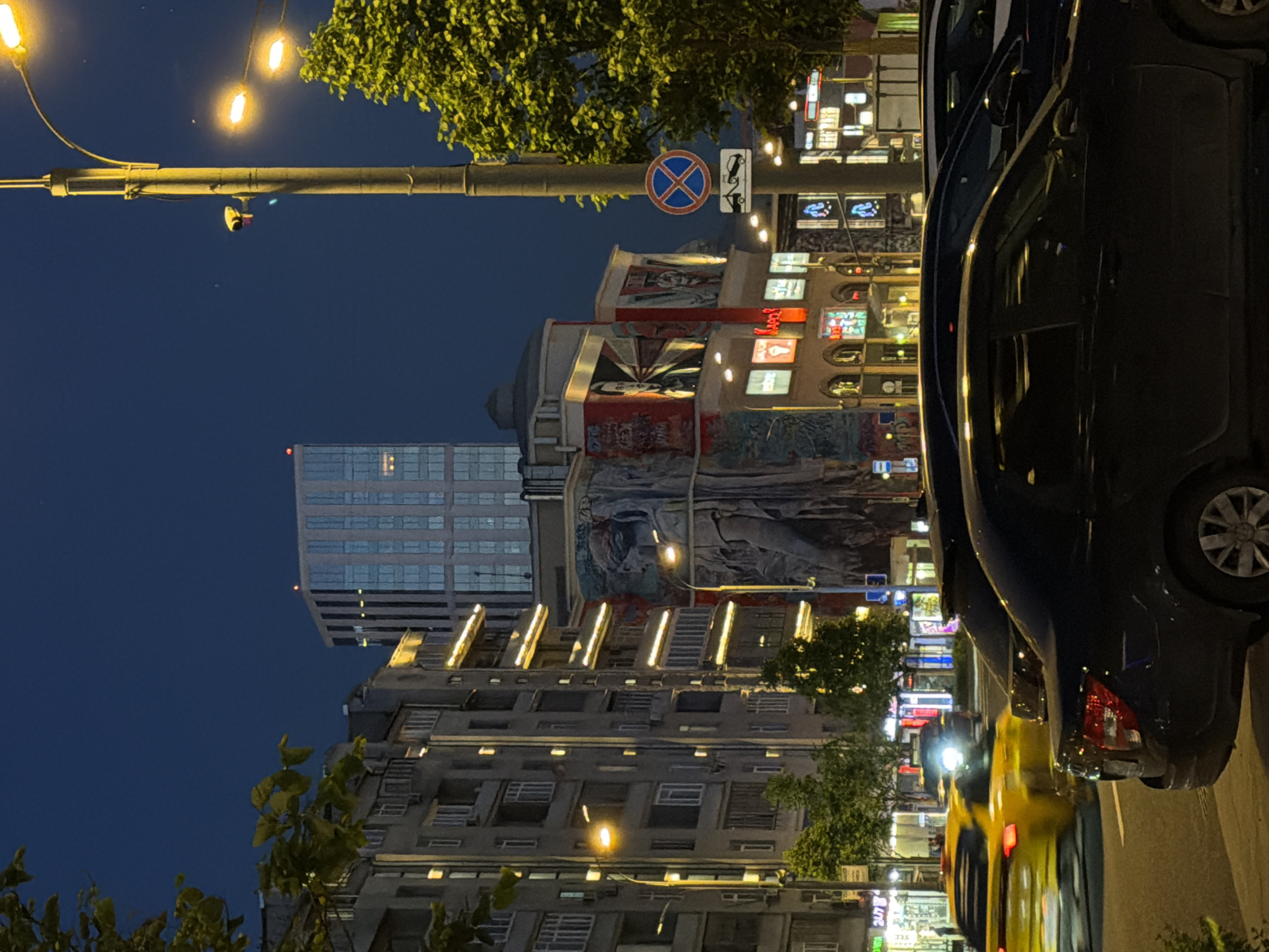The image captures a nighttime urban street scene in Moscow, Russia, specifically along Arbat Street. The prominent central building features the "Triumph of the Spirit" mural by Oleg Nikitin, depicting a large, draped classical figure predominantly in shades of blue, green, and brown. Above and to the left of this, other large murals are visible, including a stylized face of a man with an open mouth in red and black, and another figure resembling a bust in a military style.
On the left, a multi-story residential or commercial building stands, with numerous illuminated windows and horizontal light strips on some balconies. A street-level shop displays a "24/7" sign. In the background, a tall, modern high-rise building with a glass facade is illuminated, featuring a small red light at its apex.
In the midground, a street pole with several bright yellow streetlights extends across the frame. A yellow security camera is mounted on the pole, along with two traffic signs: a "No Stopping" sign (blue circle with red diagonal cross) and a "Tow Away Zone" sign below it. A tree with green foliage is visible on both the left and right sides, partially obscuring buildings and the sky.
At street level, several vehicles are present. Two dark-colored sedans are parked parallel to the curb on the right. To their left, a yellow taxi is in motion, indicated by motion blur, with its red taillights visible. Another vehicle with headlights on is faintly visible further down the street. The road surface is dark asphalt. The overall scene is characterized by the dark blue night sky, contrasting with the numerous artificial lights from streetlamps, building windows, and commercial signs. No clearly identifiable people are discernible in the immediate foreground or midground.