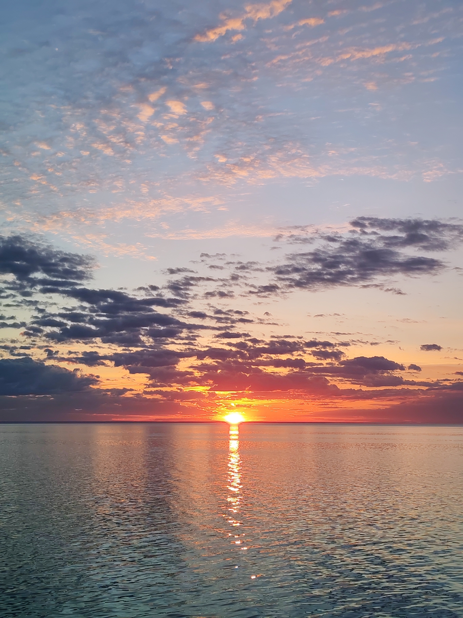 The image shows a sunset over water. The sun is setting on the horizon, casting a bright orange glow on the water and the clouds above. The sky is filled with clouds, some dark and gray, others pink and white. The water is calm and reflects the colors of the sky. The location is likely Bol'shie Mkhi, Russia, as requested, although this cannot be verified visually. There are no people visible in the image.