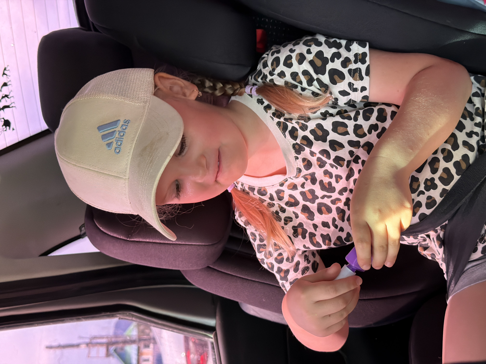A young girl is sitting in a car seat, wearing a baseball cap with the Adidas logo, and a leopard print shirt. She has blonde hair styled in braids. She is holding a small, white and purple object in her hands. The scene appears to be inside a car, with a glimpse of an outdoor environment through the window, possibly in the city of Chaikovsky, Russia.