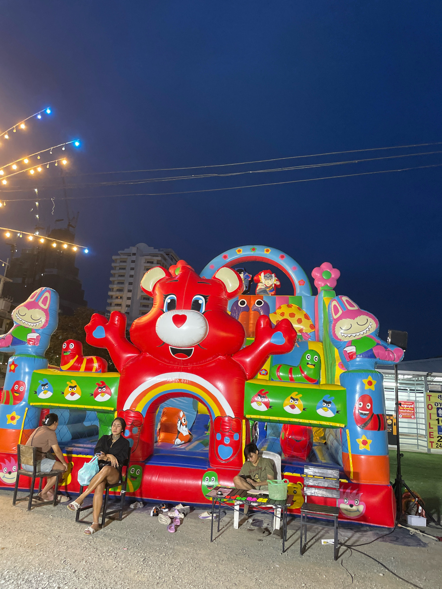 An inflatable cartoon-themed bouncy castle, featuring a large red bear character and other colorful designs, is illuminated at dusk in Pattaya City, Thailand. The bouncy castle is situated outdoors on a gravel surface, with a dark blue sky above. Several string lights are visible in the upper left portion of the image, possibly part of a street or market setup. In the foreground, two women are seated on chairs near the bouncy castle, with one woman looking at her phone. A few pairs of shoes and a bag are scattered on the ground. In the background, modern buildings and construction cranes suggest an urban environment. The scene appears to be part of an outdoor entertainment area or market in Pattaya.