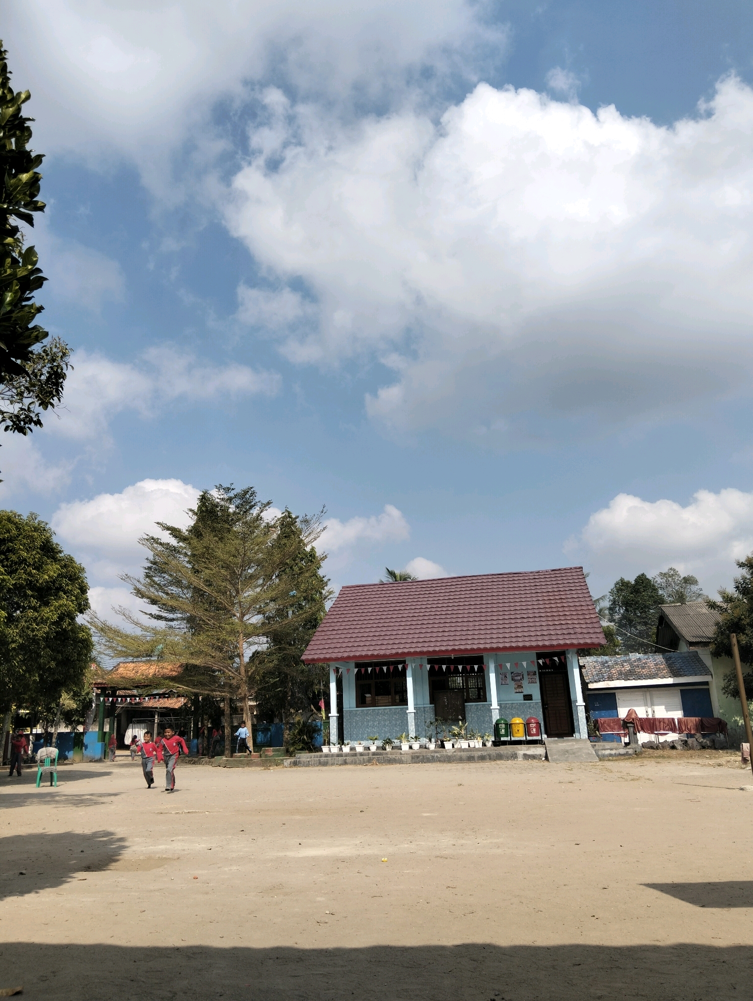 The image depicts a schoolyard in Banjar Agung, Indonesia, on a bright, cloudy day. In the foreground, a sandy, open space is visible, with shadows cast from unseen objects to the right and left. Two young boys in matching red and grey uniforms are running towards the left of the frame. Behind them, a building with a reddish-brown tiled roof and light blue walls stands prominently. This building has a covered porch with potted plants and banners hanging along the railing. To the left of this main building, another structure with a terracotta-tiled roof is partially visible, suggesting a larger school complex. Several trees with green foliage are scattered throughout the scene, with one particularly tall, slender tree with sparse leaves standing near the center. The sky is a vibrant blue, filled with fluffy white clouds. The overall atmosphere is that of a typical school day in a rural or semi-urban Indonesian setting.