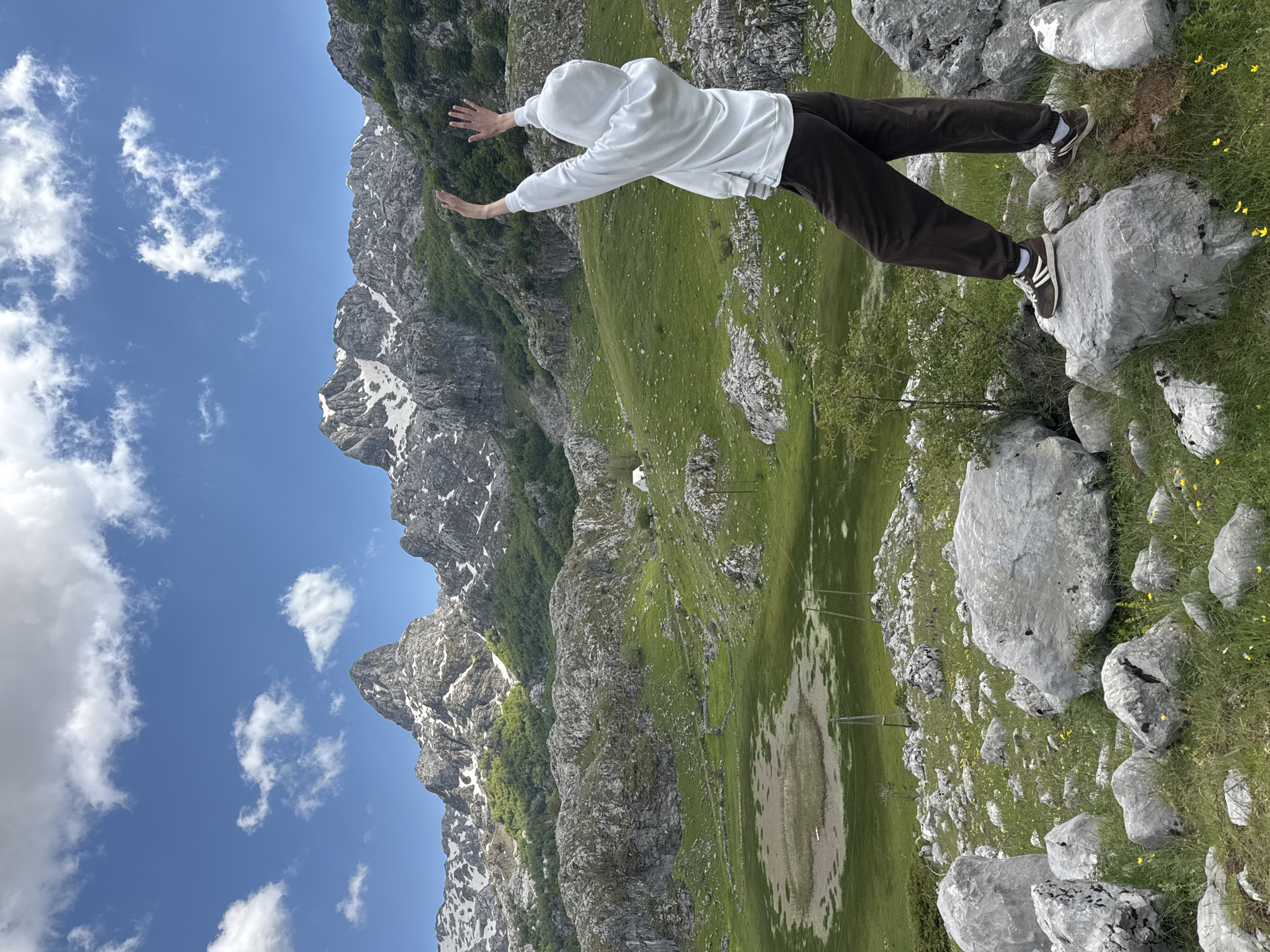 A person in a white hooded sweatshirt and brown pants is standing on a large rock outcropping. The person's arms are outstretched. The background features a landscape of grassy hills, rocky terrain, and mountains with patches of snow. In the distance, power lines run across a meadow-like area that could be prone to flooding based on ground texture and coloration. The scene is Brskut, Montenegro.