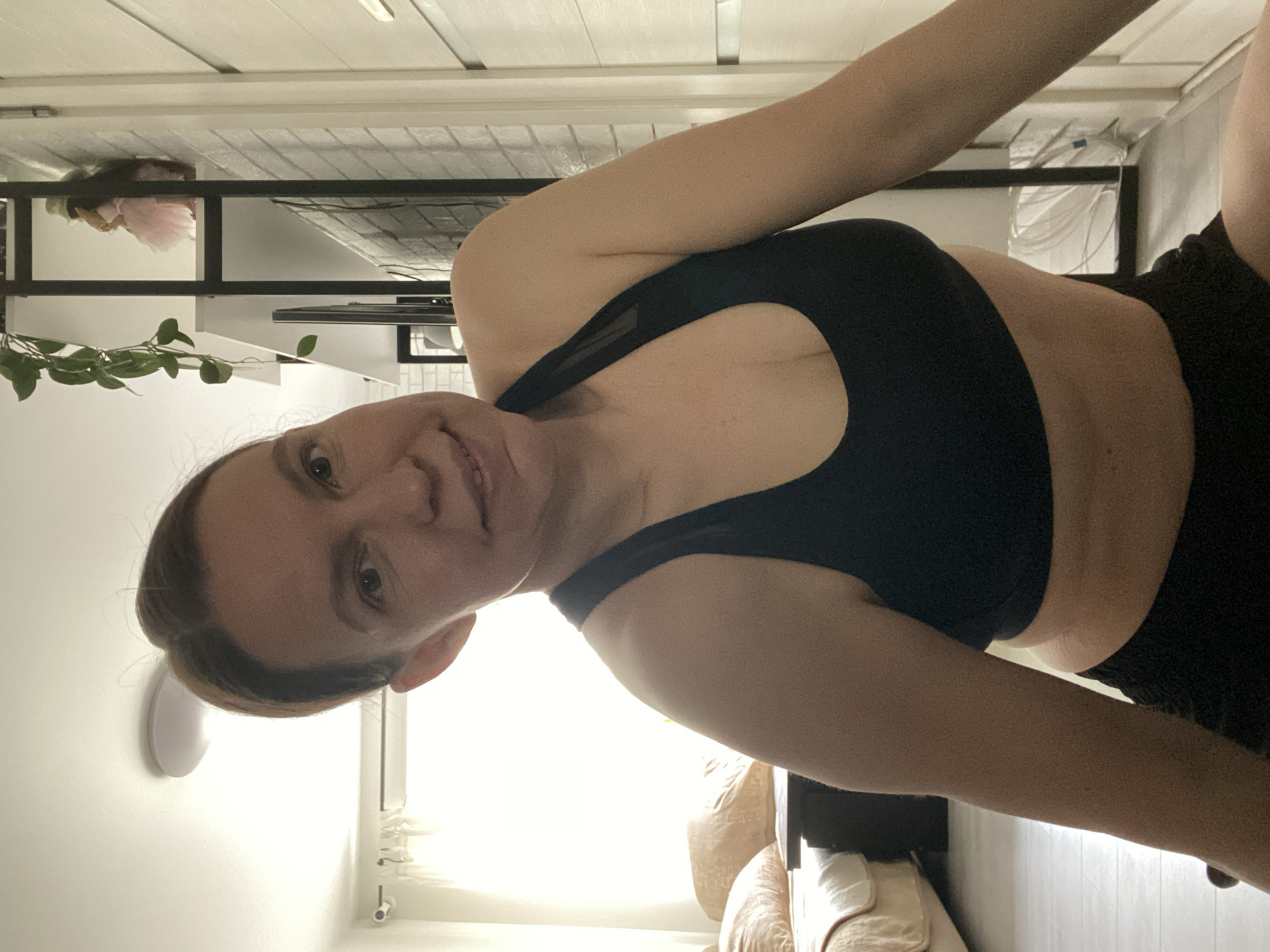 A woman is shown in an indoor setting, wearing a black sports bra and black shorts. She is positioned facing the camera. The location appears to be a domestic interior, possibly an apartment. The background includes a white wall, a light fixture, a black shelf unit, and a glimpse of what appears to be a sofa or cushioned chair. There is no information to indicate the location is Chaikovsky, Russia.