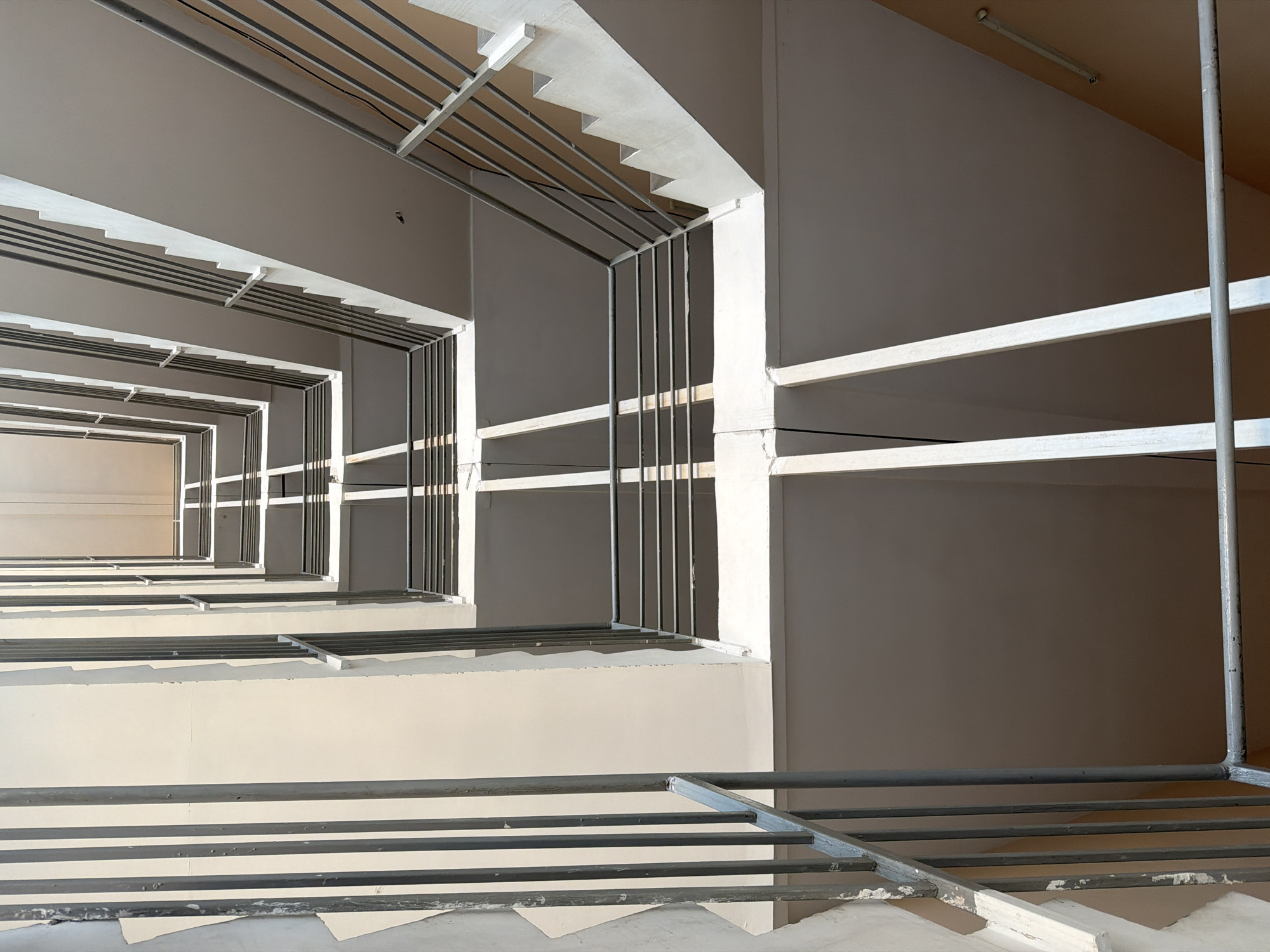 The image depicts an upward view of a multi-story building's open-air stairwell. The architecture features concrete floors and walls with metal railings and support beams. The staircases are visible winding upwards, with light casting sharp shadows across the surfaces. The scene suggests an interior or semi-enclosed space within a building, possibly a residential complex or an office building. There are no people visible in the image. The overall impression is one of architectural geometry and the interplay of light and shadow. There is no information in the image to suggest a specific location in Kyiv, Ukraine.