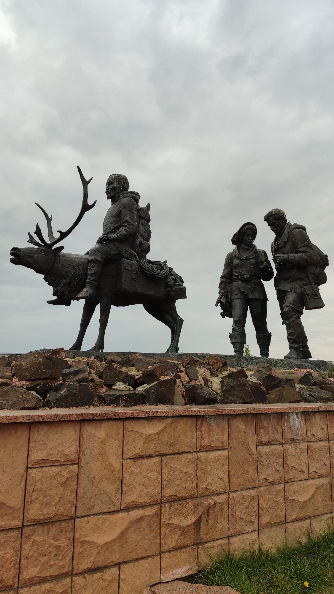The image shows a bronze monument depicting three figures and a reindeer. In the foreground, a stone wall made of roughly hewn tan blocks supports a rocky foundation. On top of the rocks are three bronze figures. The leftmost figure is seated on a reindeer, which is facing left. The rider is wearing a parka and backpack, and the reindeer is laden with luggage. To the right of the reindeer rider, two more figures stand side-by-side, appearing to be in conversation. The figure on the left is a woman wearing a parka and holding a rifle. The figure on the right is a man, also in outdoor clothing, looking at the woman and gesturing. The monument is set against a backdrop of a cloudy, overcast sky. This monument is located in Mirny, Russia, and it represents the explorers and indigenous peoples who have inhabited and traveled through the region.