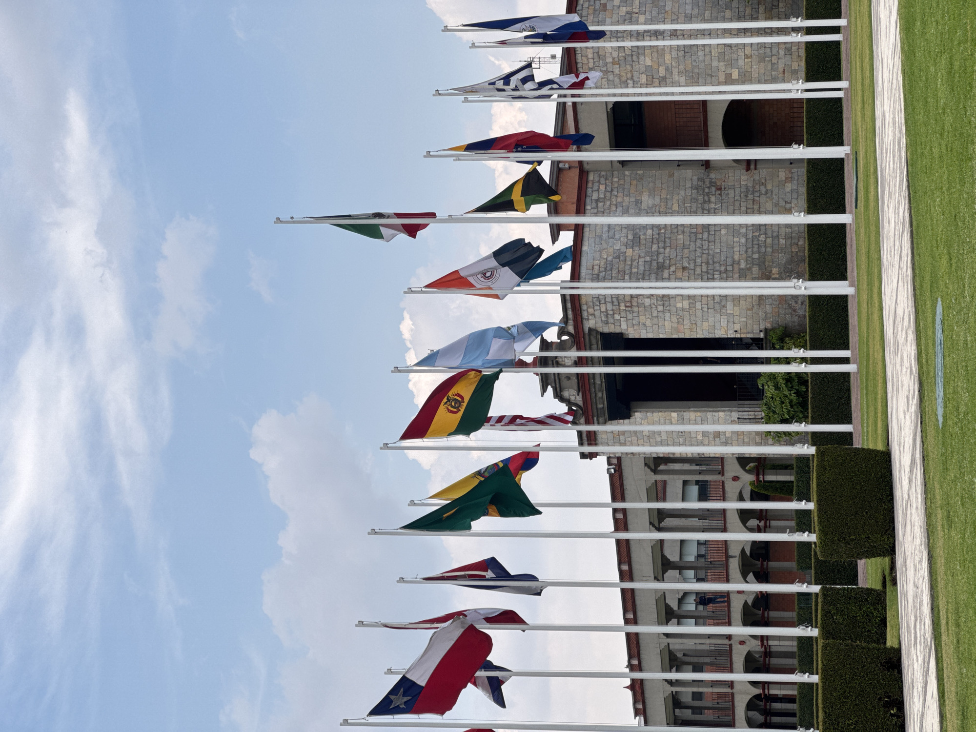 The original image displays a vibrant outdoor scene captured in a portrait orientation, featuring a long line of international flags set against a bright, partly cloudy blue sky. Due to the image's rotation, the flagpoles extend horizontally across the frame, and the flags appear to hang either upwards or downwards.
From bottom to top in the image, the visible flags include Peru, Chile, Panama, Brazil, Bolivia, Argentina, the Organization of American States (OAS), Italy, Jamaica, Venezuela, Greece, and El Salvador. This collection suggests a gathering of nations, particularly with a strong representation from the Americas, alongside European flags.
Behind the flag display, a stately building with a light-colored stone or brick facade is visible. It features multiple dark, arched openings and sections of red-tiled roofing or trim. In front of the building, a meticulously trimmed dark green hedge runs parallel to the flagpoles. To the right of the flagpoles, a wide, well-maintained green lawn stretches out, bisected by a light-colored paved pathway. The overall setting is formal and well-kept, typical of a public institution or diplomatic grounds.
The bright lighting indicates it is daytime with clear weather. No people or visible text are present in the image. The location is San Andrés Cholula, Mexico.